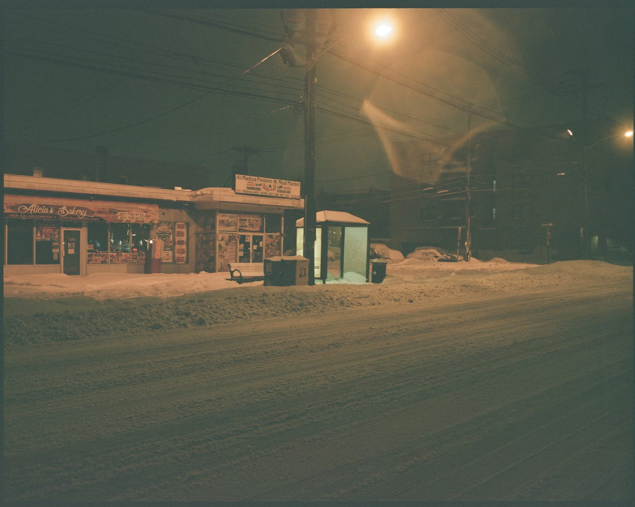 A snow-covered street at night with a bakery storefront, bus stop, and parked cars illuminated by streetlights.