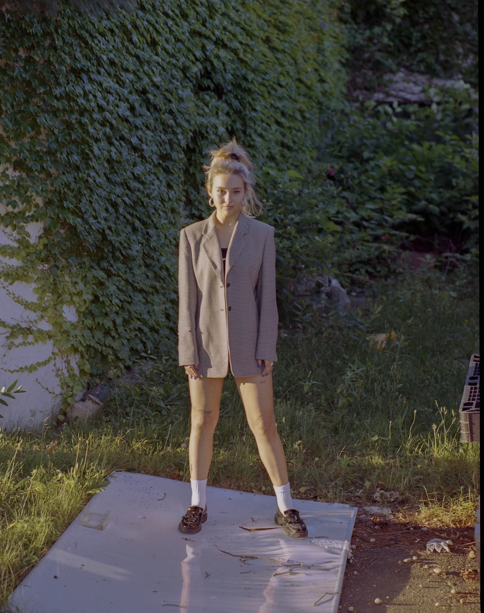 A young woman standing outdoors on a white platform with grass and green bushes in the background. She is wearing a beige blazer, shorts, white socks, and sneakers, with her hair tied up in a messy bun.