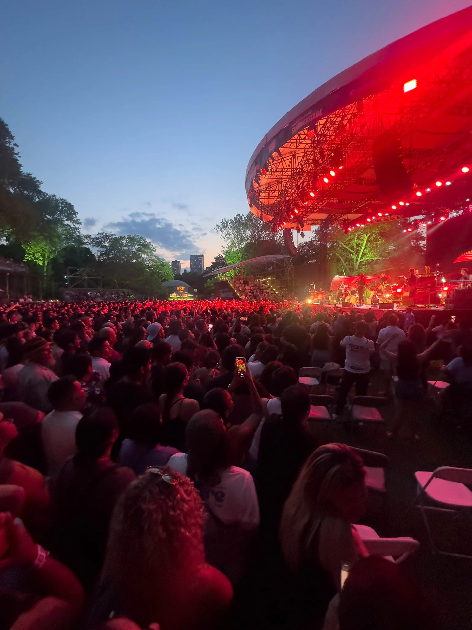 Crowd watching a concert at an outdoor amphitheater during twilight, with stage lights illuminating the stage.
