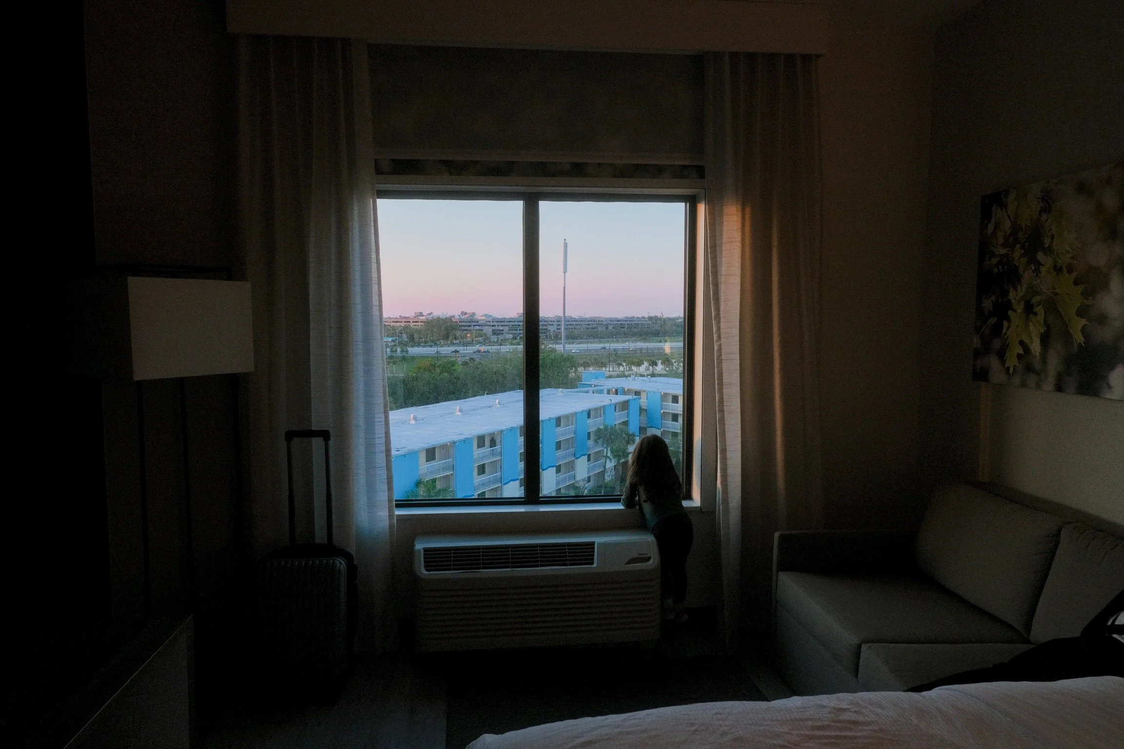 A young girl standing by a hotel room window, looking outside at the cityscape during dusk or dawn, with the sky showing pastel colors and the buildings and greenery visible outside.