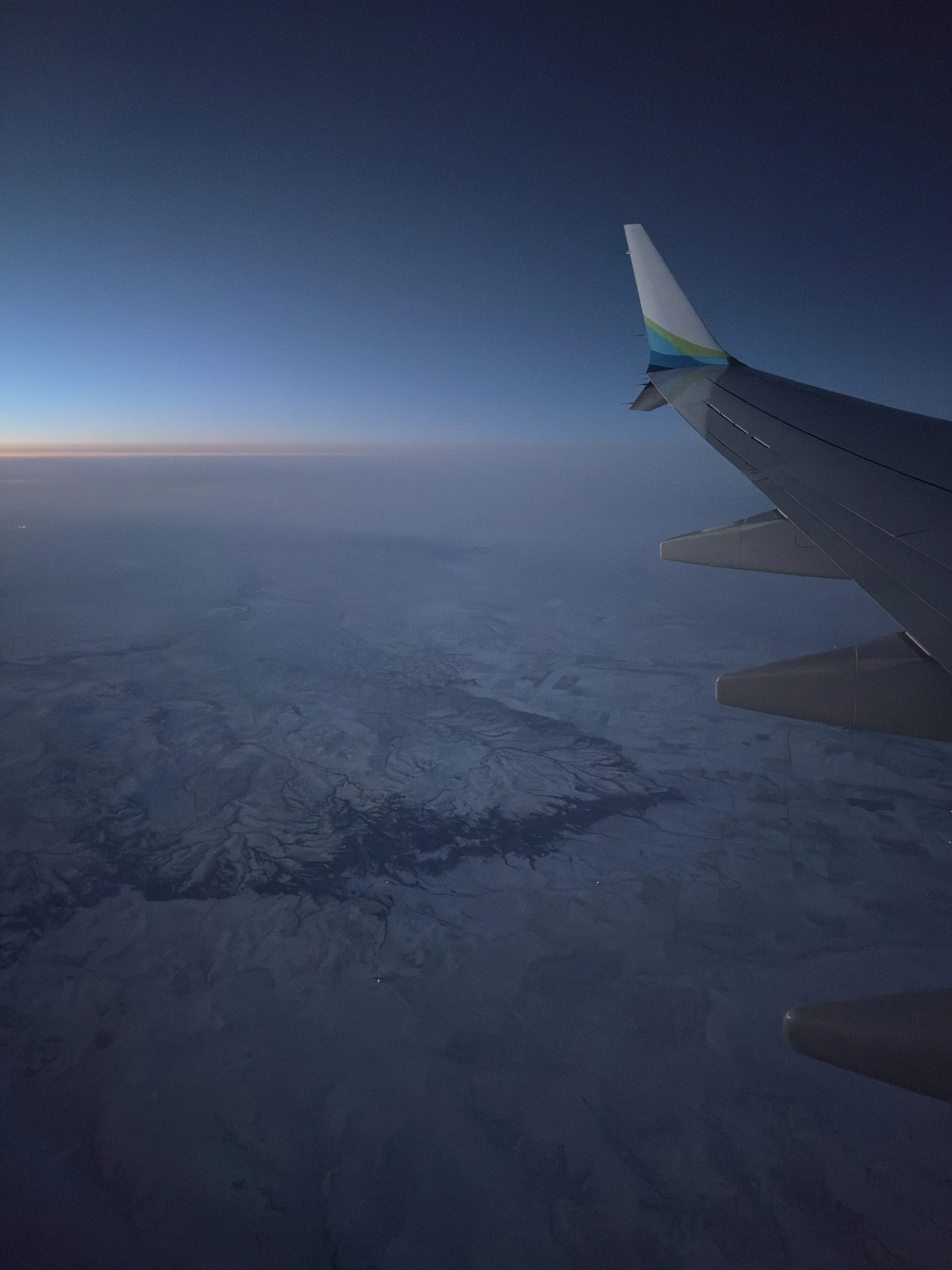 View from an airplane window showing the wing against a twilight sky with a snow-covered landscape below.