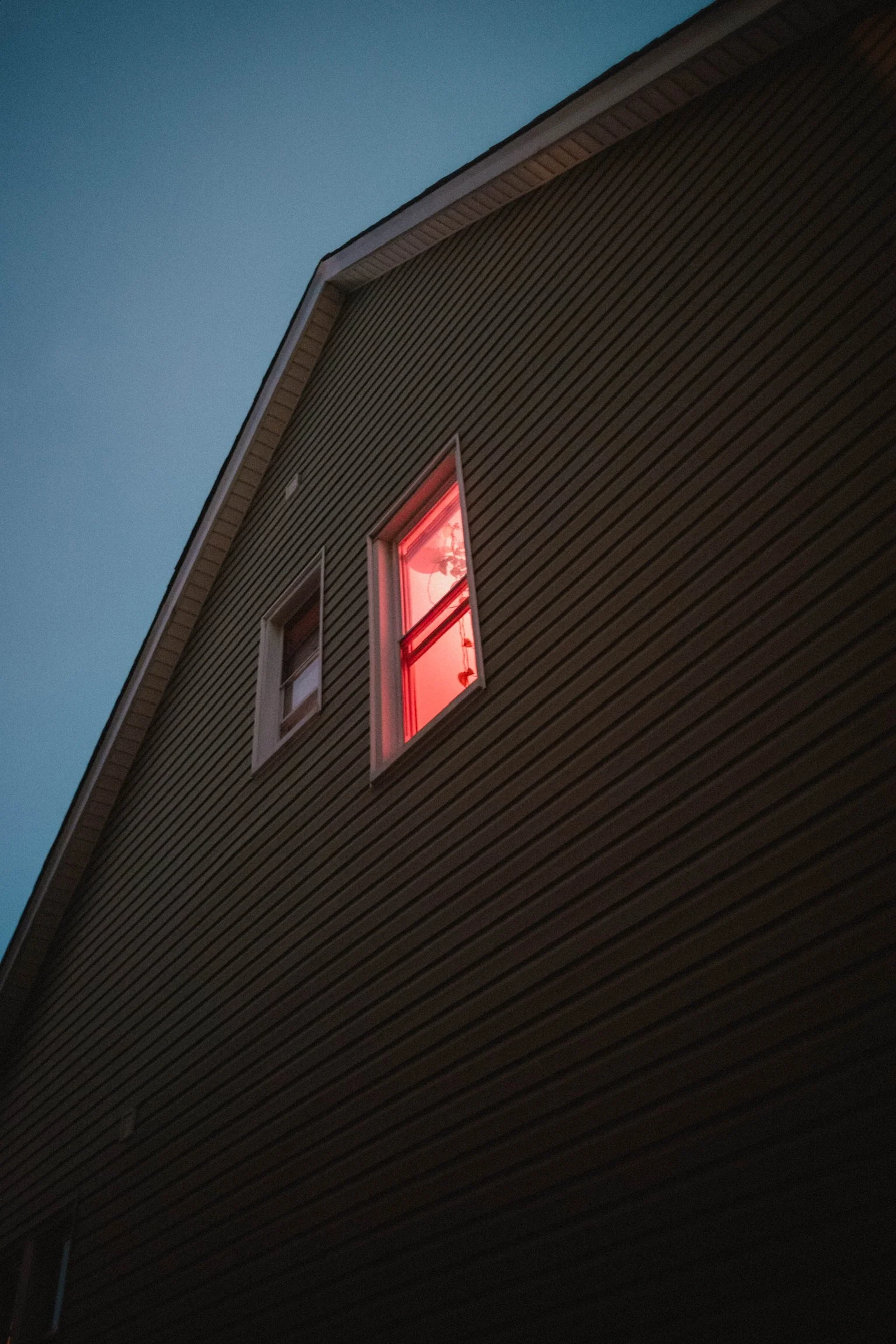 A house's exterior at night with two windows; one window emits a red glow, suggesting a light inside, while the other window is dark. The house has brown horizontal siding and a light-colored trim.