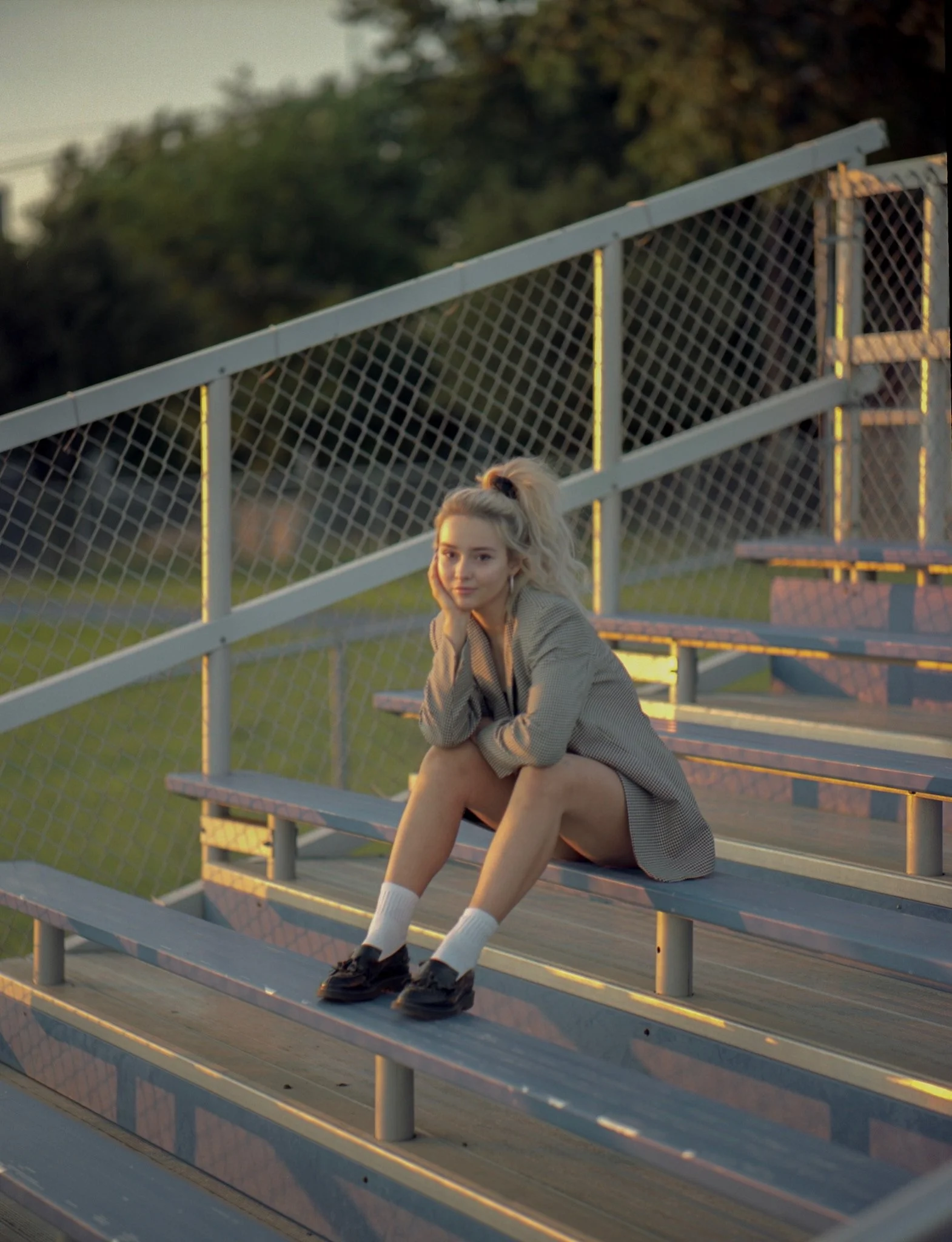 A young woman with blonde hair tied in a high ponytail, sitting on metal bleachers at a sports field during sunset, looking at the camera with a relaxed expression.