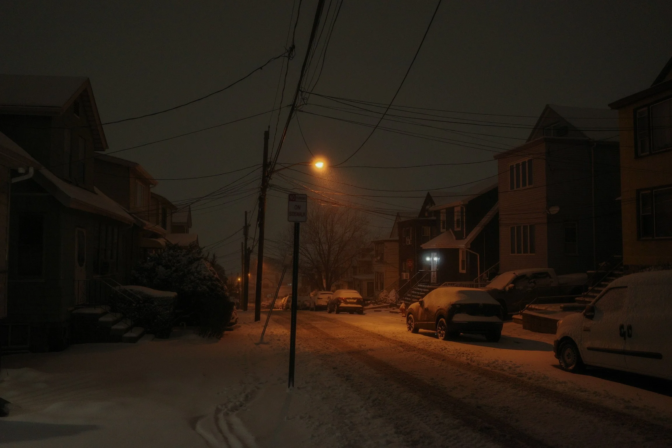 Snow-covered residential street at night with parked cars, houses, streetlights, utility poles, and overhead wires in a winter scene.