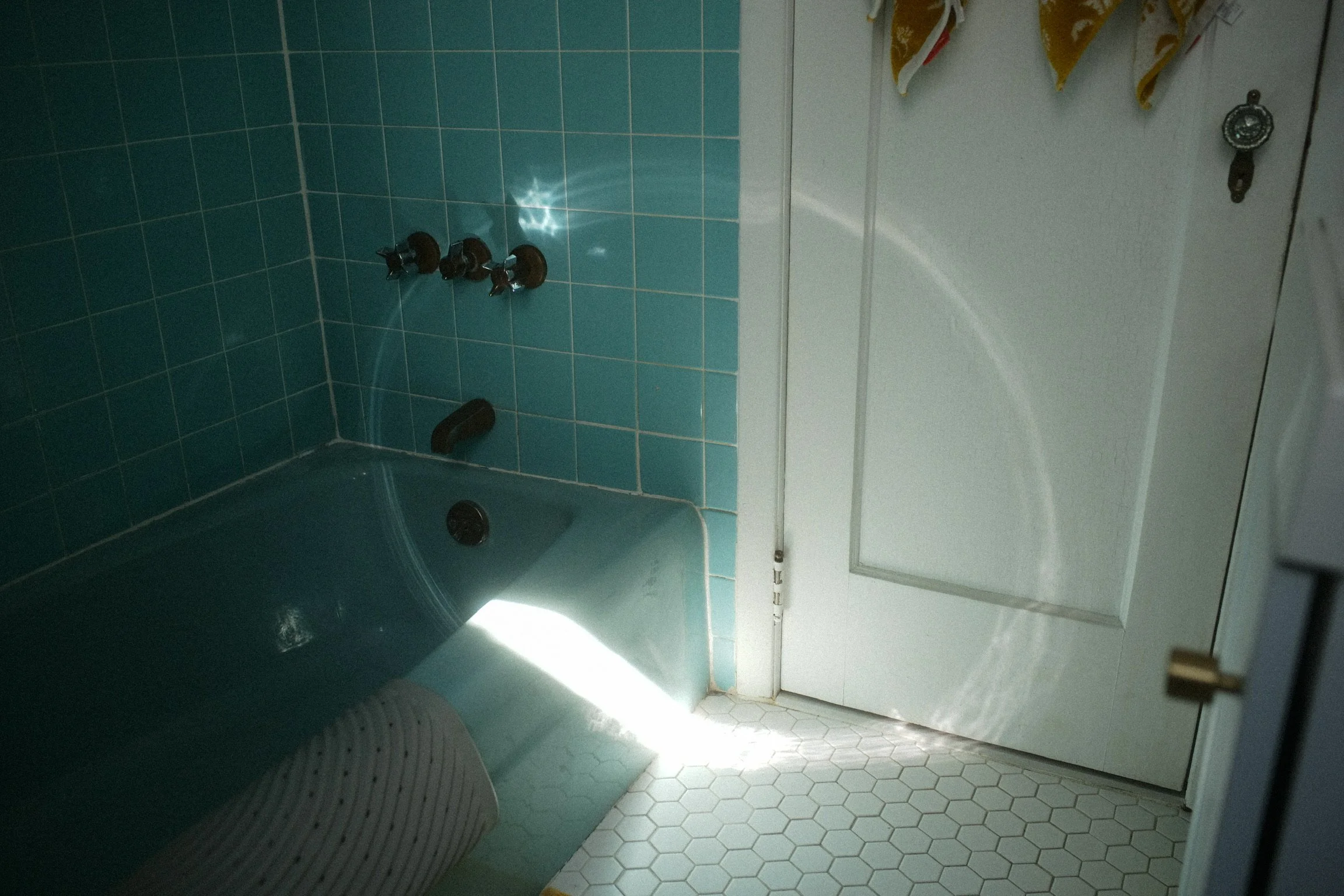 A bathroom with teal wall tiles, a blue bathtub, three black faucet handles, and a white door with a revolving lock; hexagonal floor tiles with a small patch of bright sunlight.