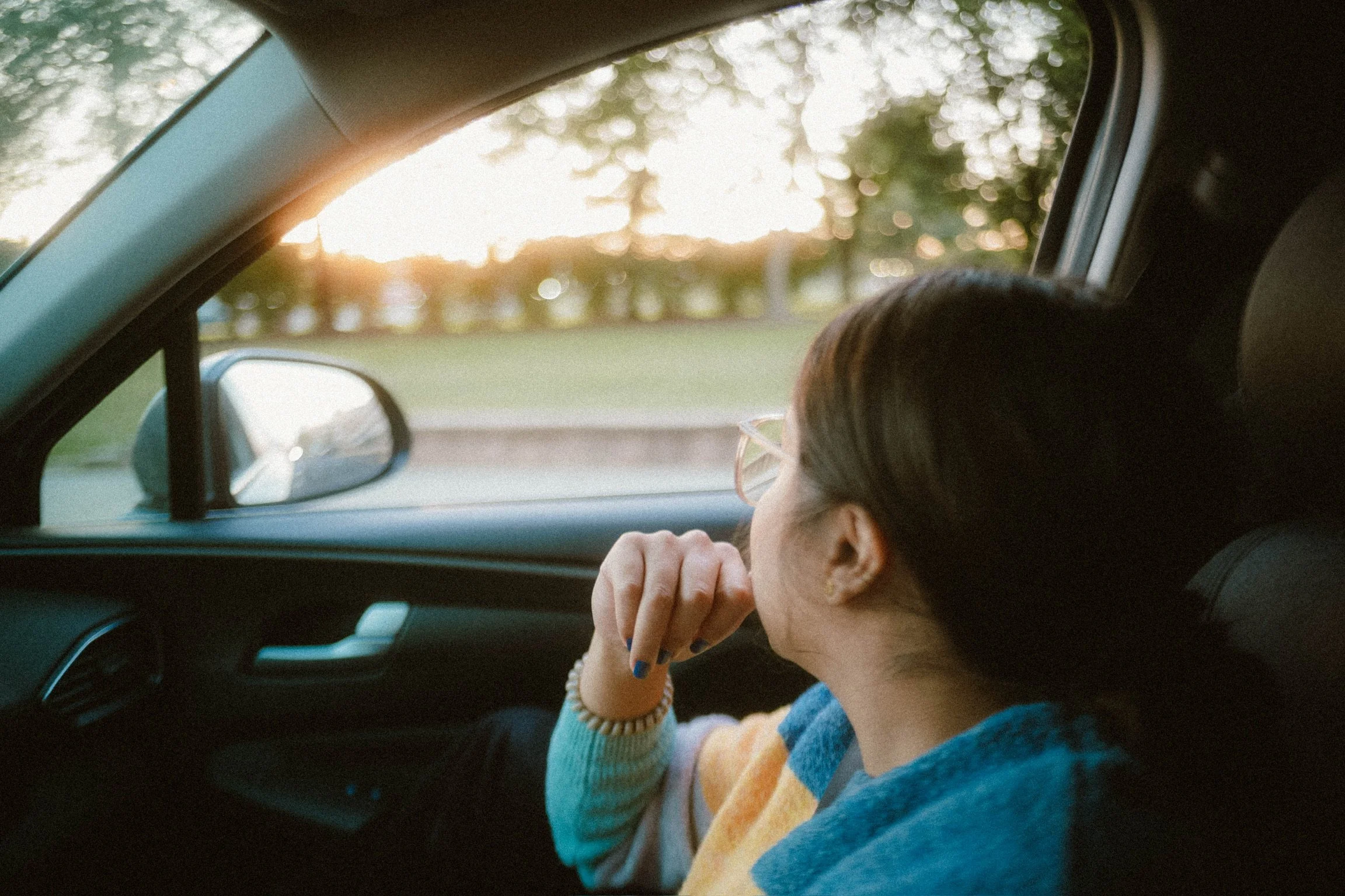 A woman sitting in the passenger seat of a car, looking out the window at a sunset over trees.
