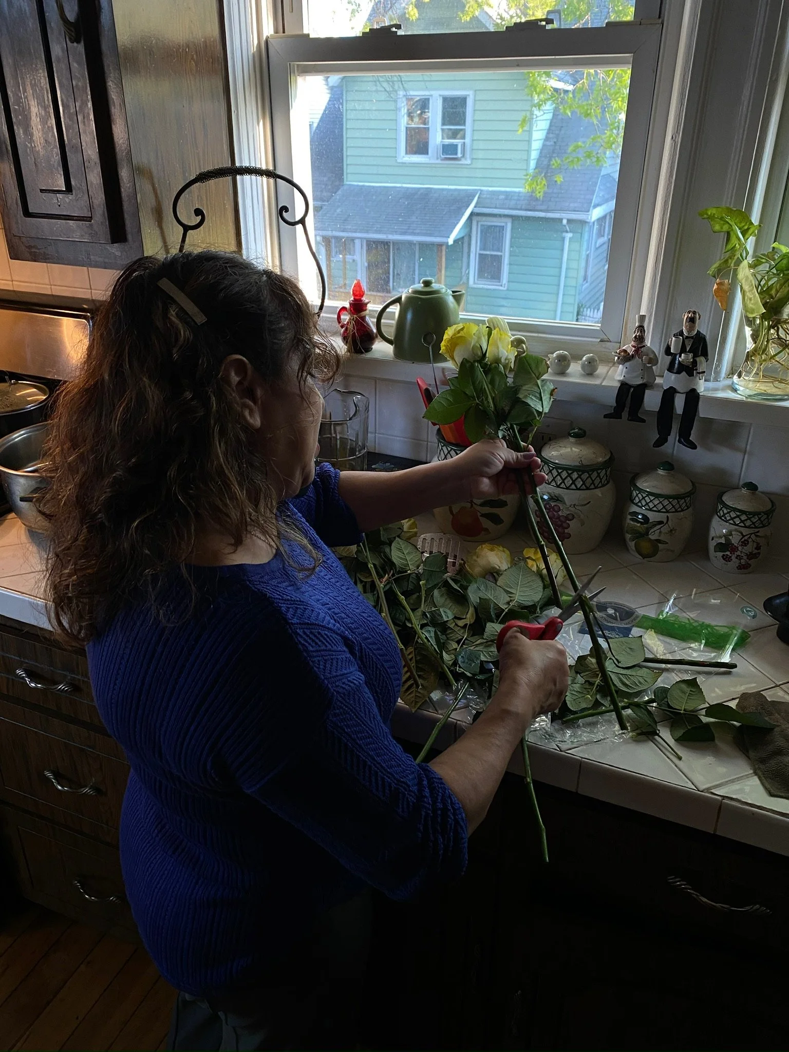 A woman in a blue sweater standing at a kitchen counter, cutting flowers with pruning shears. The counter has various items including leaves, stems, and flower arrangements. The kitchen window behind her shows neighboring houses.