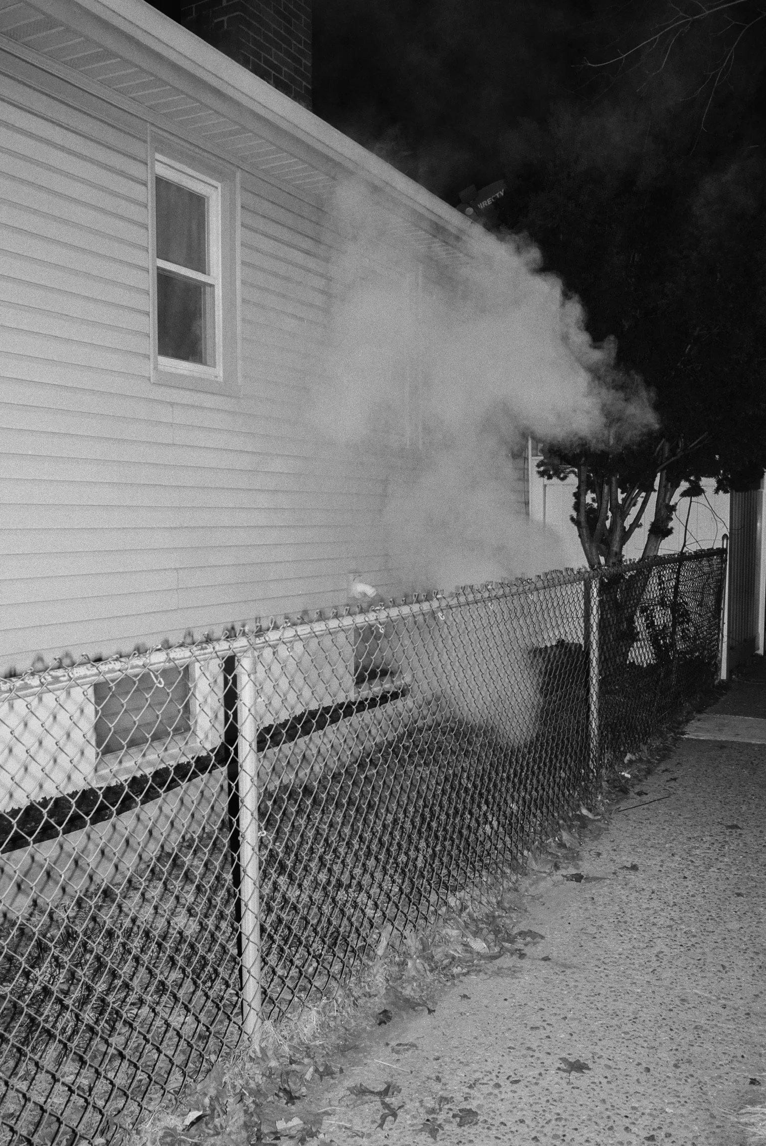 A house with siding exterior at night emitting smoke or steam through a vent or crack near the window, with a chain-link fence and walkway in the foreground.