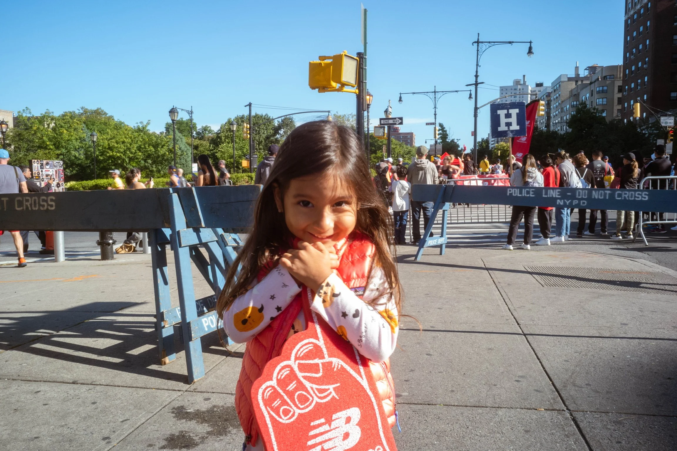 A young girl smiling at the camera, holding a red New Balance bag, standing in front of a barricade at a street event with many people, city buildings, and trees in the background.