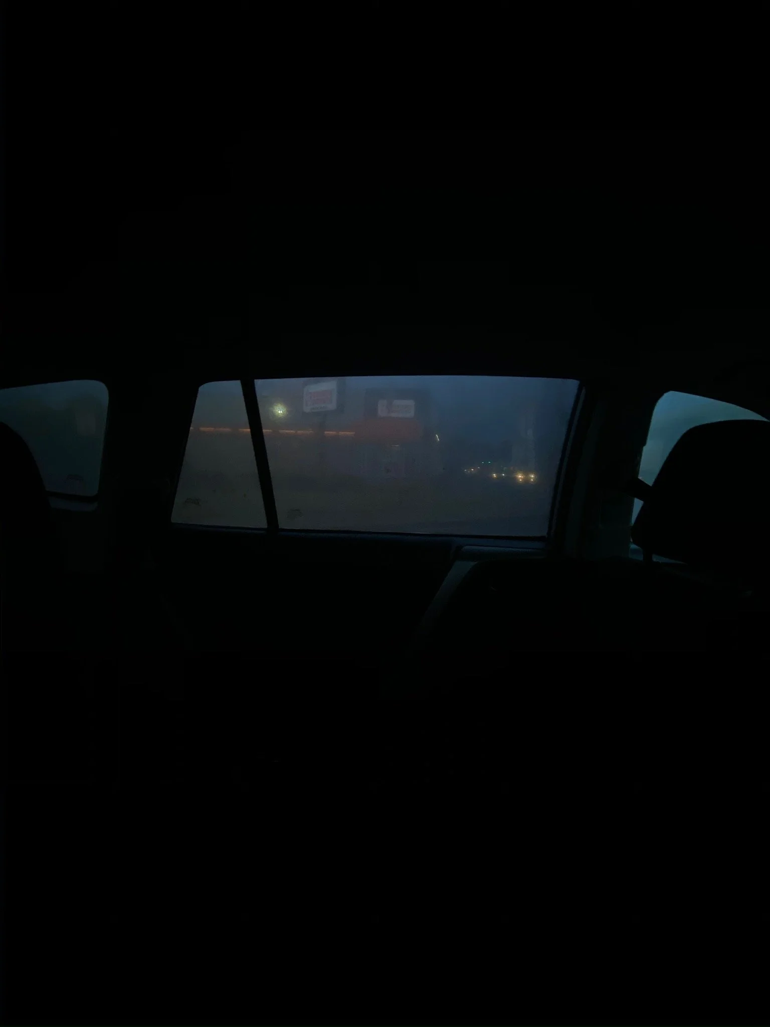 Inside a car at dusk, view through the rear window showing blurry city lights in the distance.