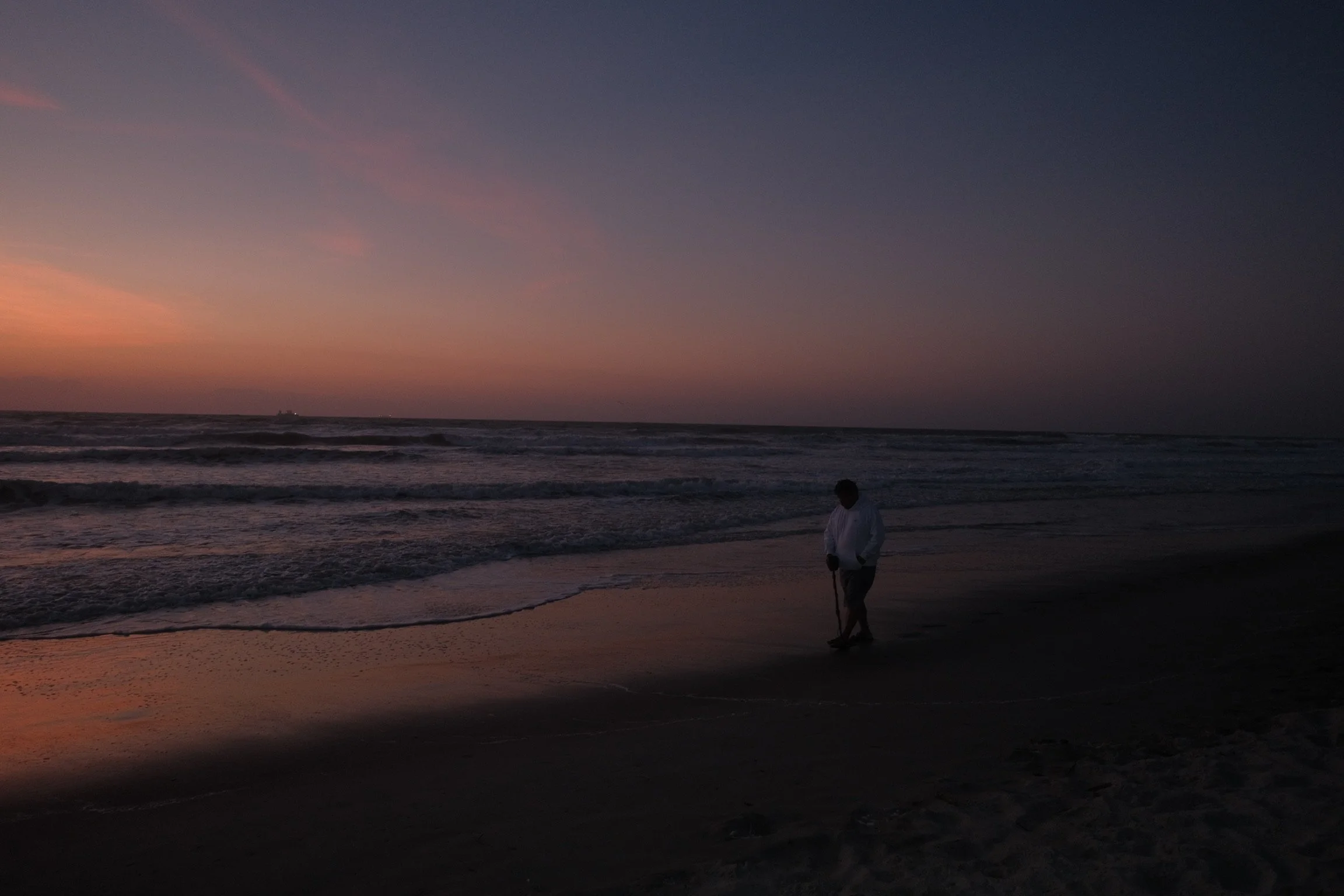 Person walking along the beach at sunset, with the ocean waves and a ship on the horizon.