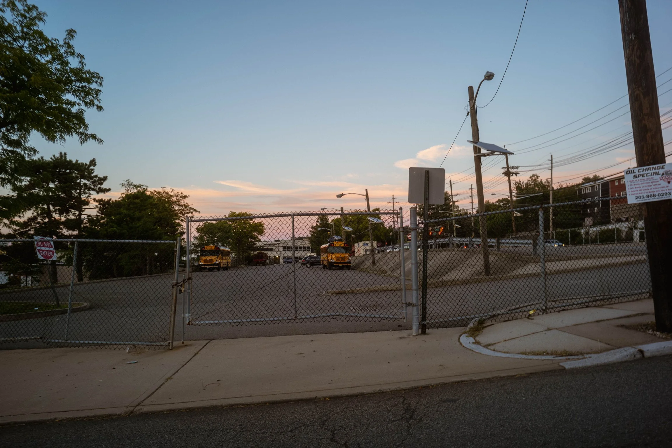 An empty parking lot surrounded by a chain-link fence with a gate, with parked yellow school buses and trees in the background, under a partly cloudy sky at sunset.