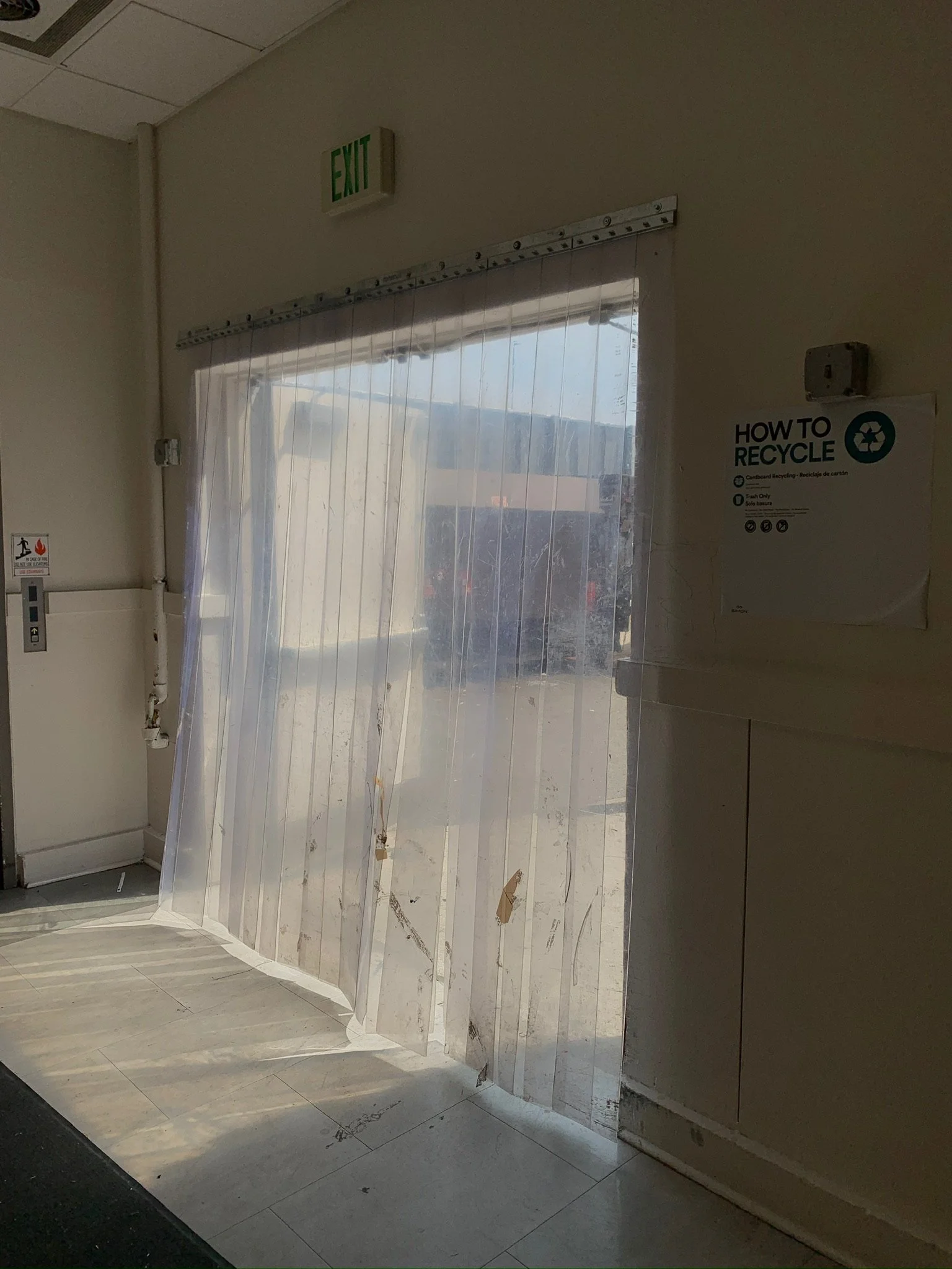 A store or office entrance with a clear plastic curtain, an illuminated green 'EXIT' sign above, and a 'HOW TO RECYCLE' sign on the wall to the right.