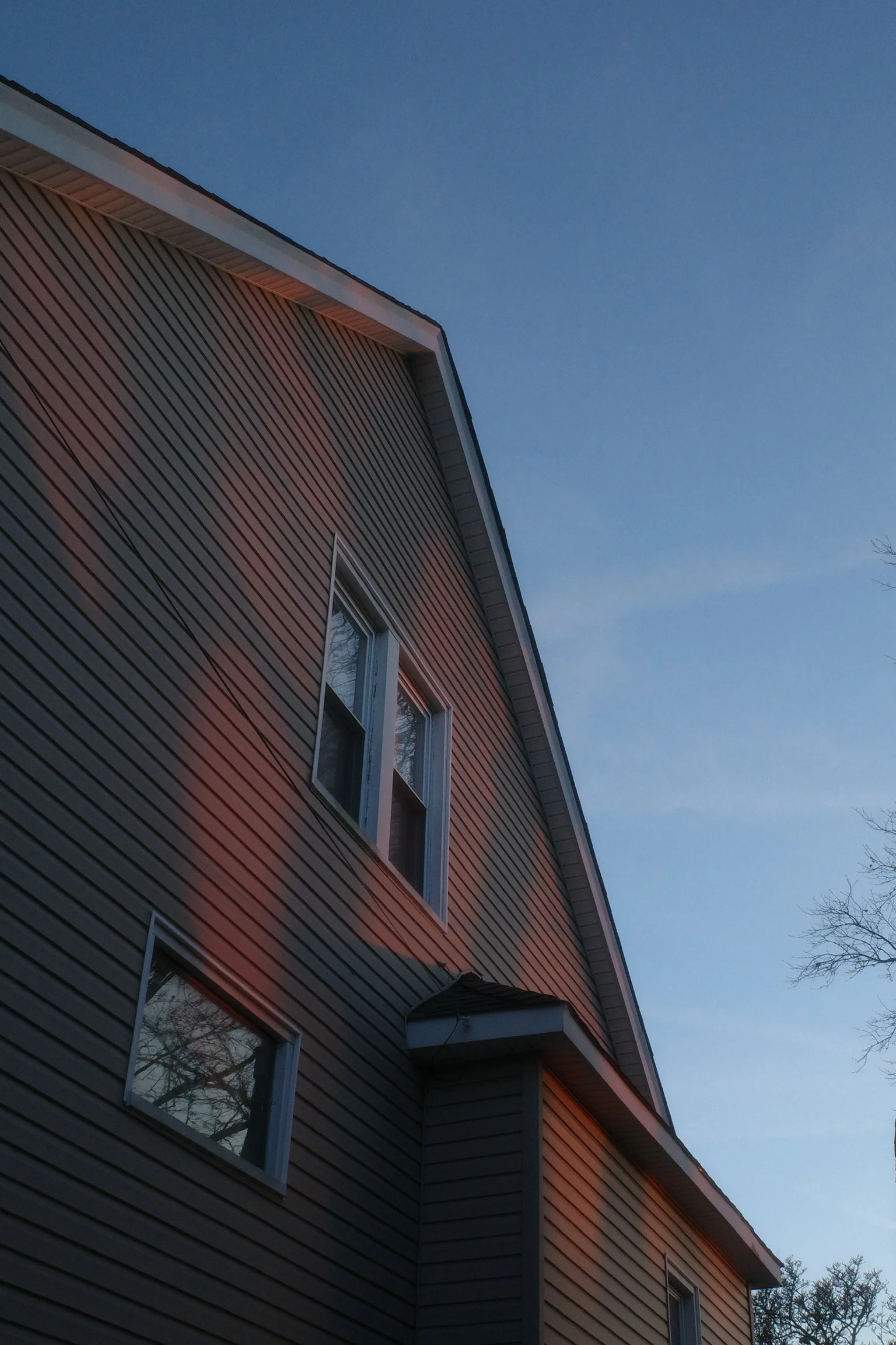 A house with beige vinyl siding and a gable roof, with sunset light casting a reddish glow on the upper part of the house, against a blue sky.