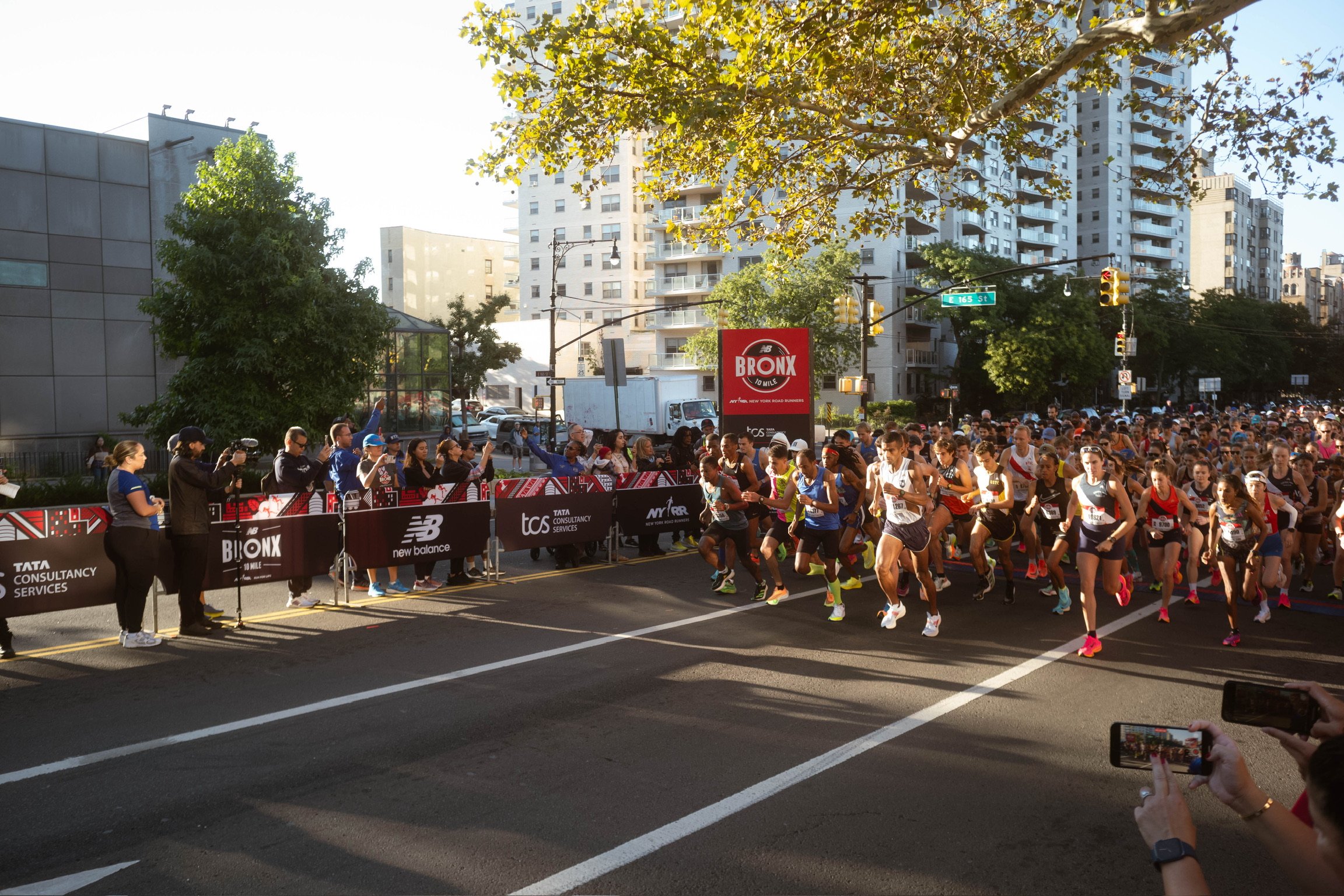 A large group of runners participating in a marathon or race in an urban setting, crossing the starting line, with spectators and photographers on the sides, trees, buildings, and traffic lights in the background.
