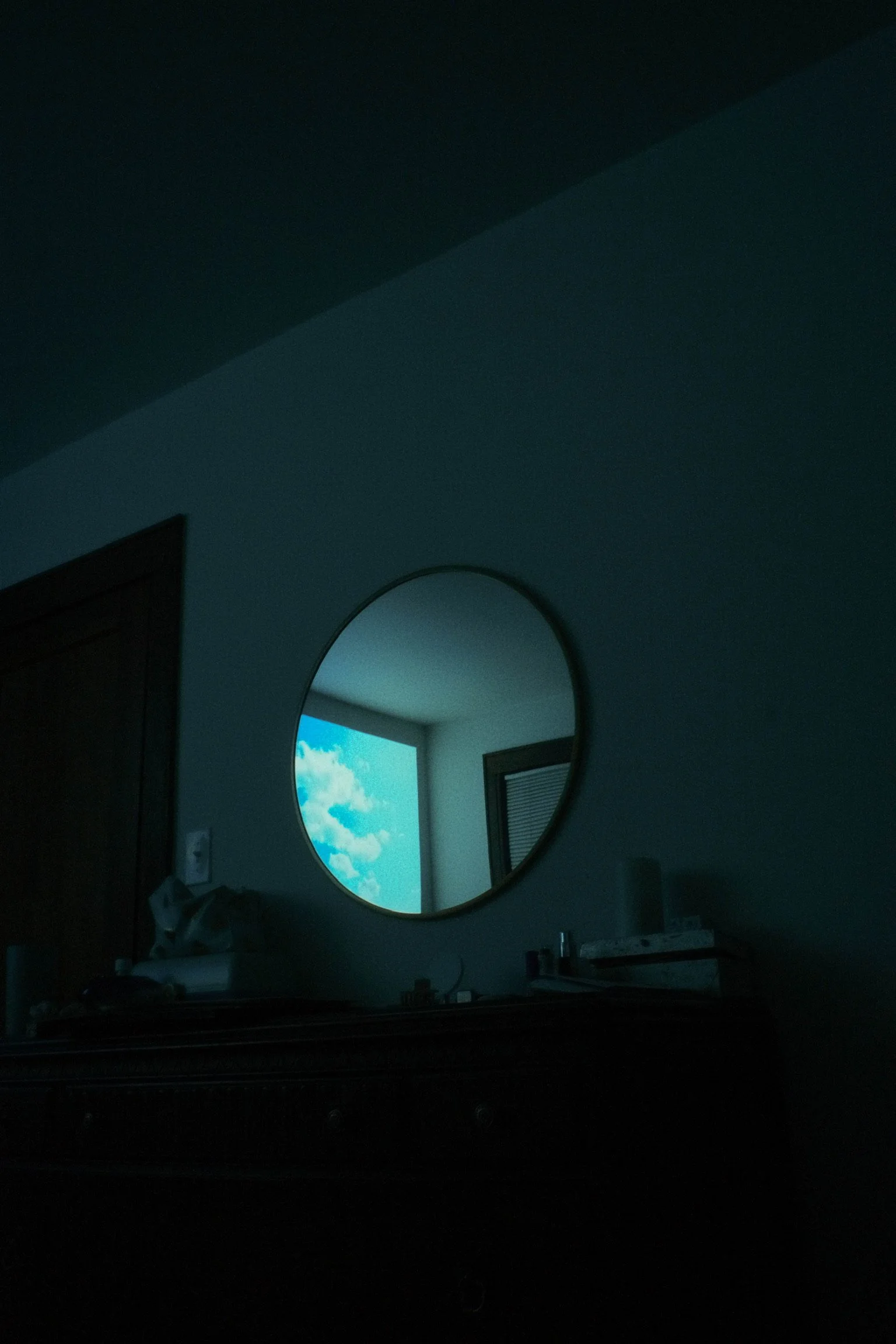 Reflection of blue sky with clouds in a round mirror above a dark dresser in a dimly lit room.