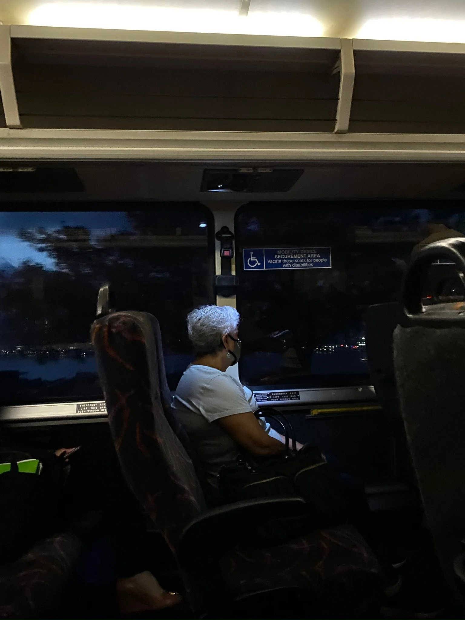 An elderly woman with gray hair and a black face mask, sitting on a bus seat, looking out the window at night. She has a black handbag on her lap, and there are luggage bags on the seat next to her.