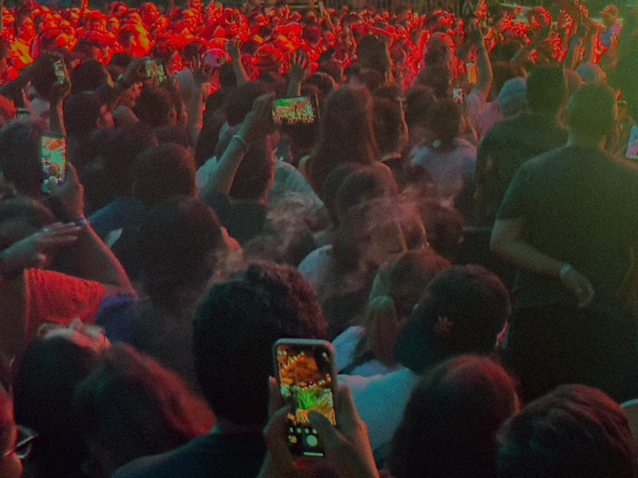 Crowd of people at a concert or event, many are taking photos or videos with their smartphones, illuminated by red and green stage lighting.