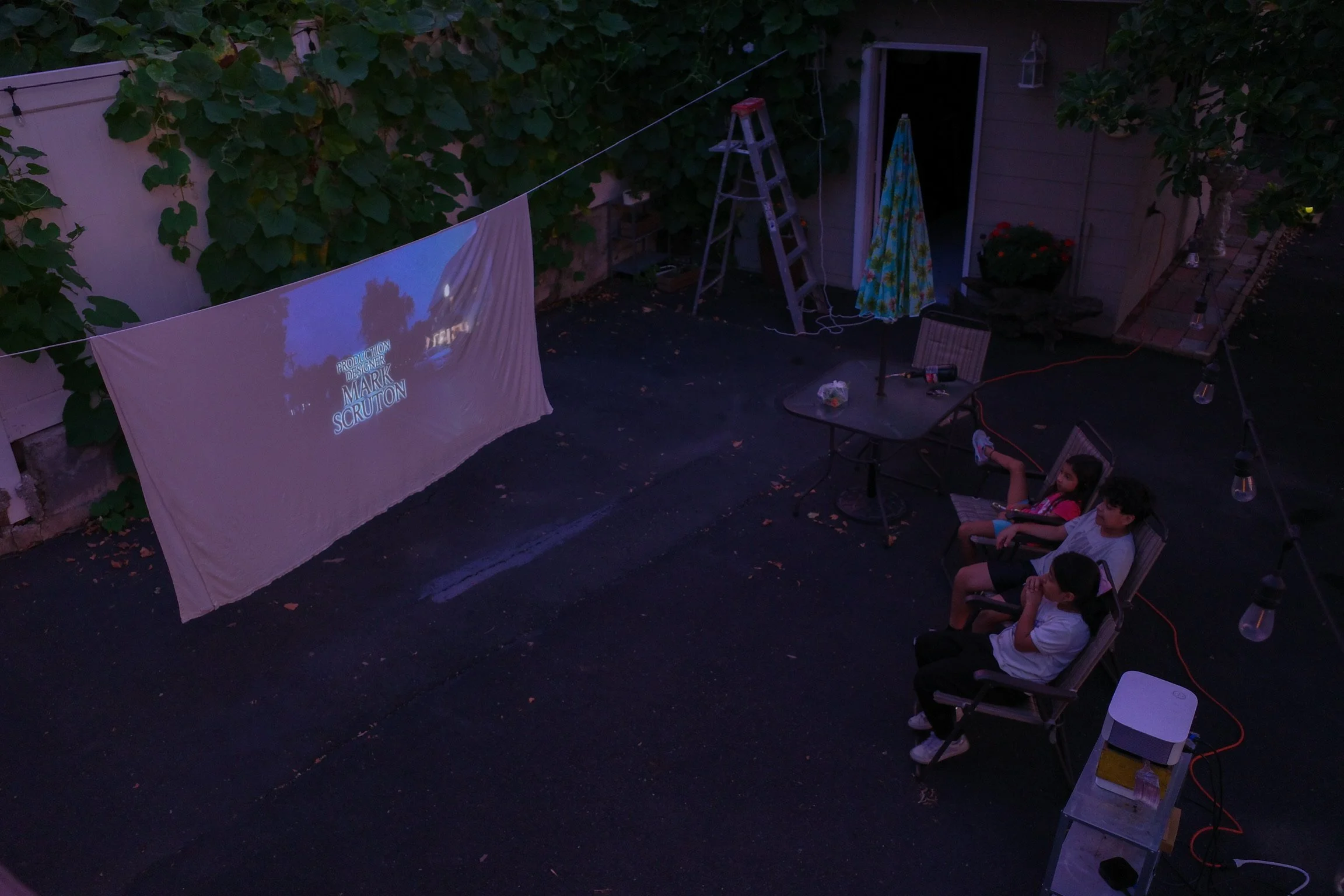 Children sitting in outdoor chairs watching a movie projected on a sheet hanging on a clothesline at dusk.