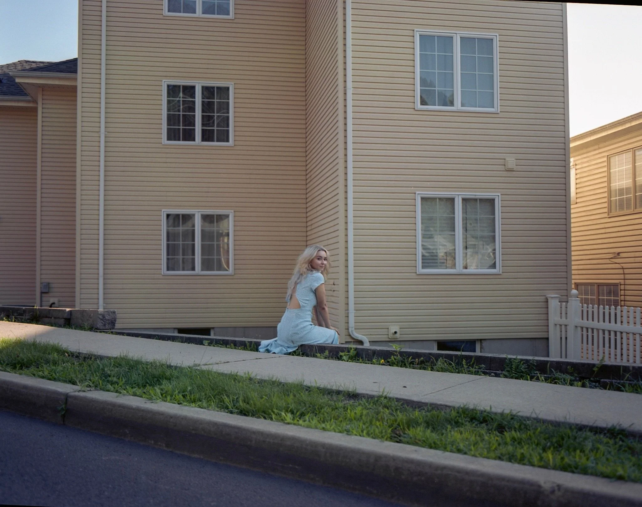 A woman with blonde hair in a light blue dress sitting on a concrete sidewalk beside a yellow building with multiple windows.