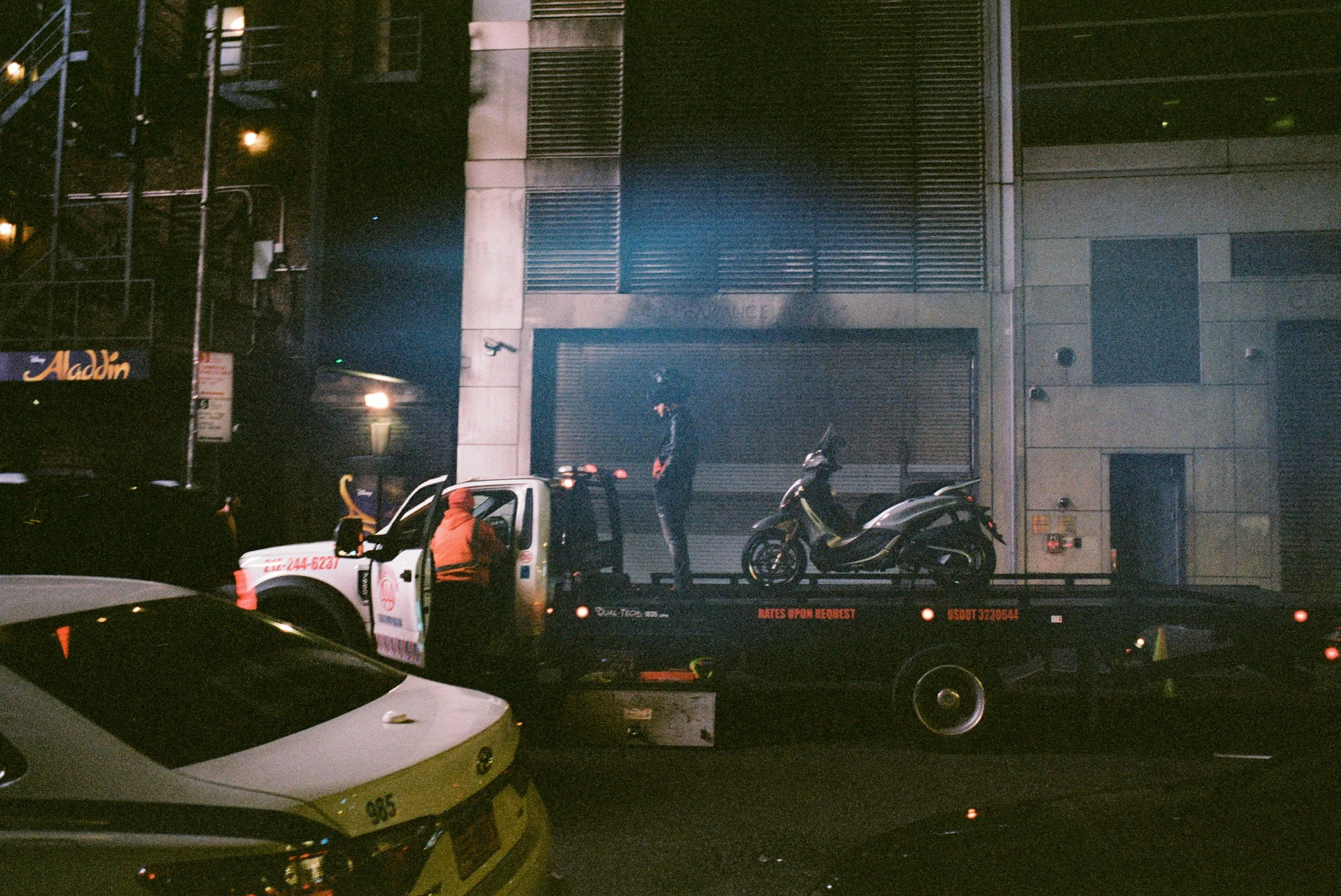 Nighttime scene showing a tow truck carrying a motorcycle and a person standing nearby, with another person in a hooded jacket talking to someone in the truck, and a motorcycle parked on the truck bed against a building with closed shutters.