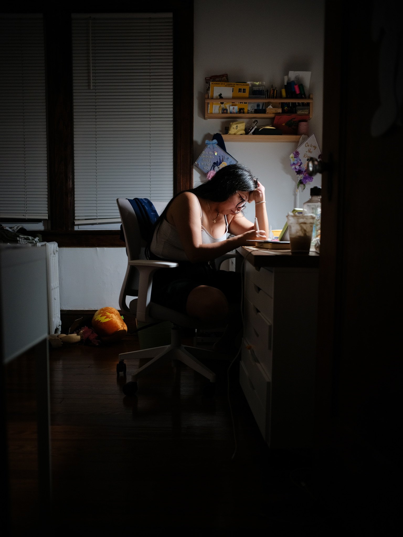 A woman sitting at a desk, studying or working, with a flashlight or lamp illuminating her workspace in a dim room. There are decorations and items on the wall behind her, and a stuffed toy on the floor nearby.