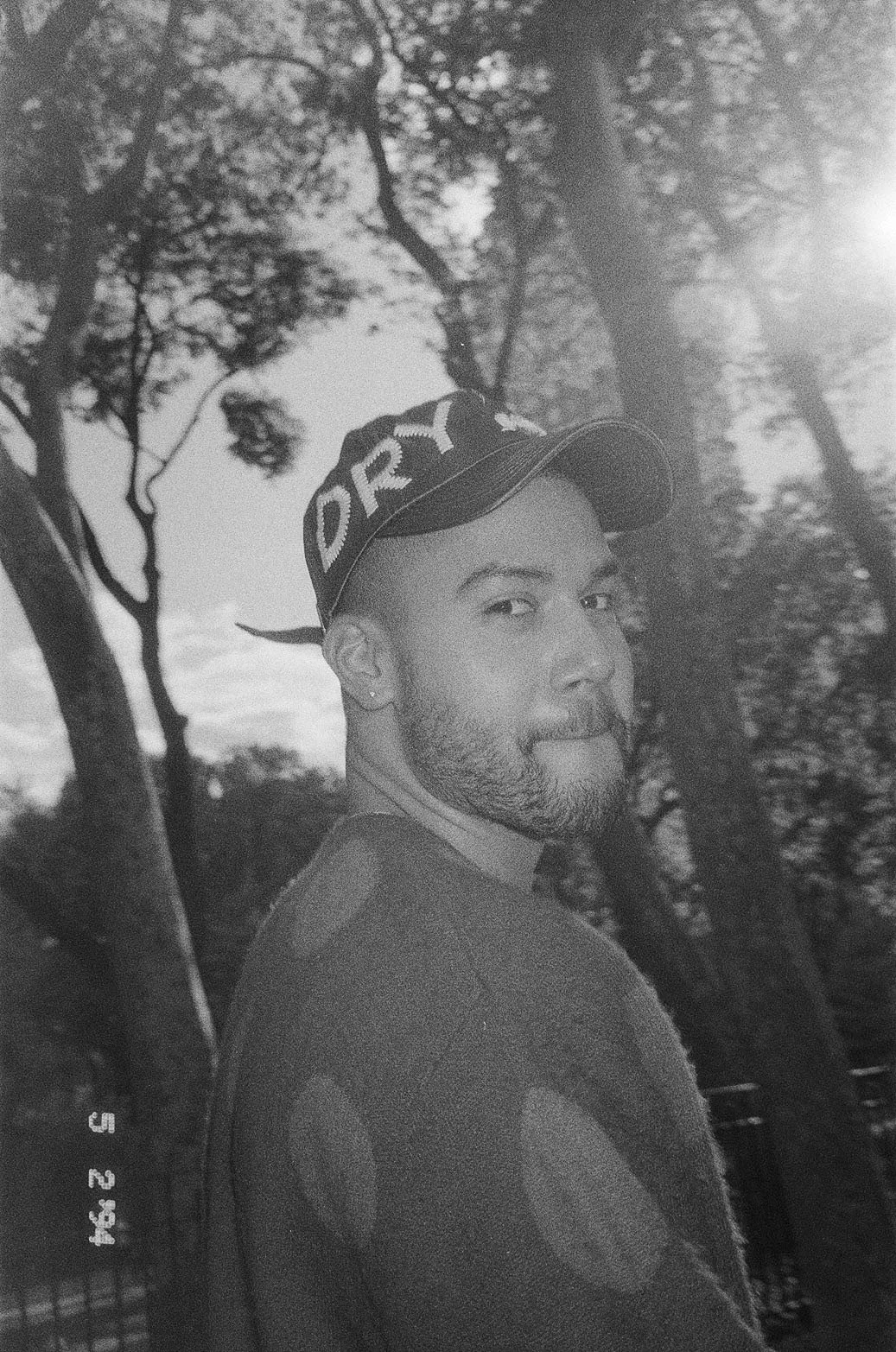 A black and white photo of a man with a beard and earrings, looking over his shoulder at the camera. He is wearing a cap with the word 'DORT' visible on it and a long sleeve shirt, with trees in the background.