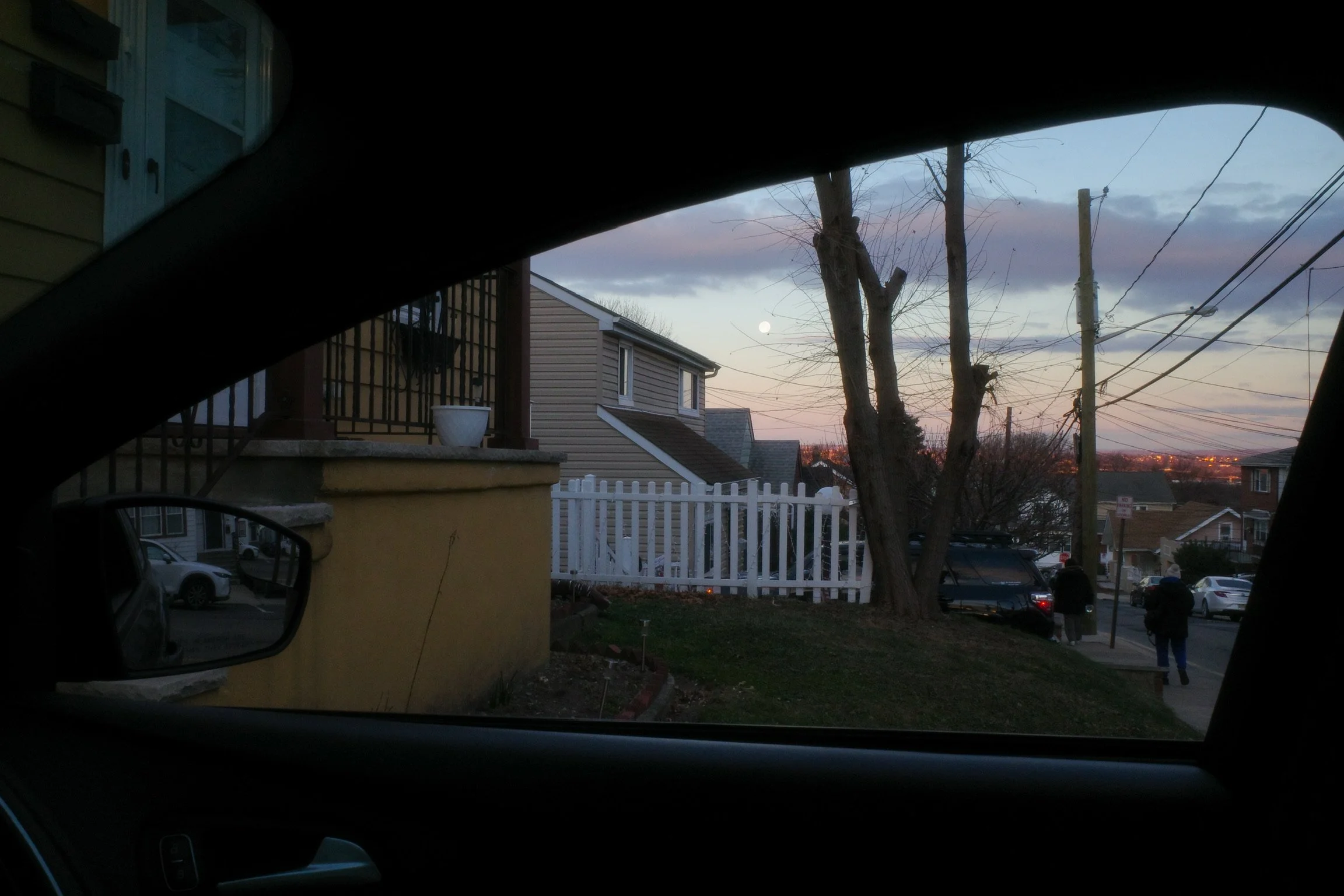 View of a residential neighborhood at dusk seen from inside a car, including houses, a tree, power lines, and the moon in the sky.