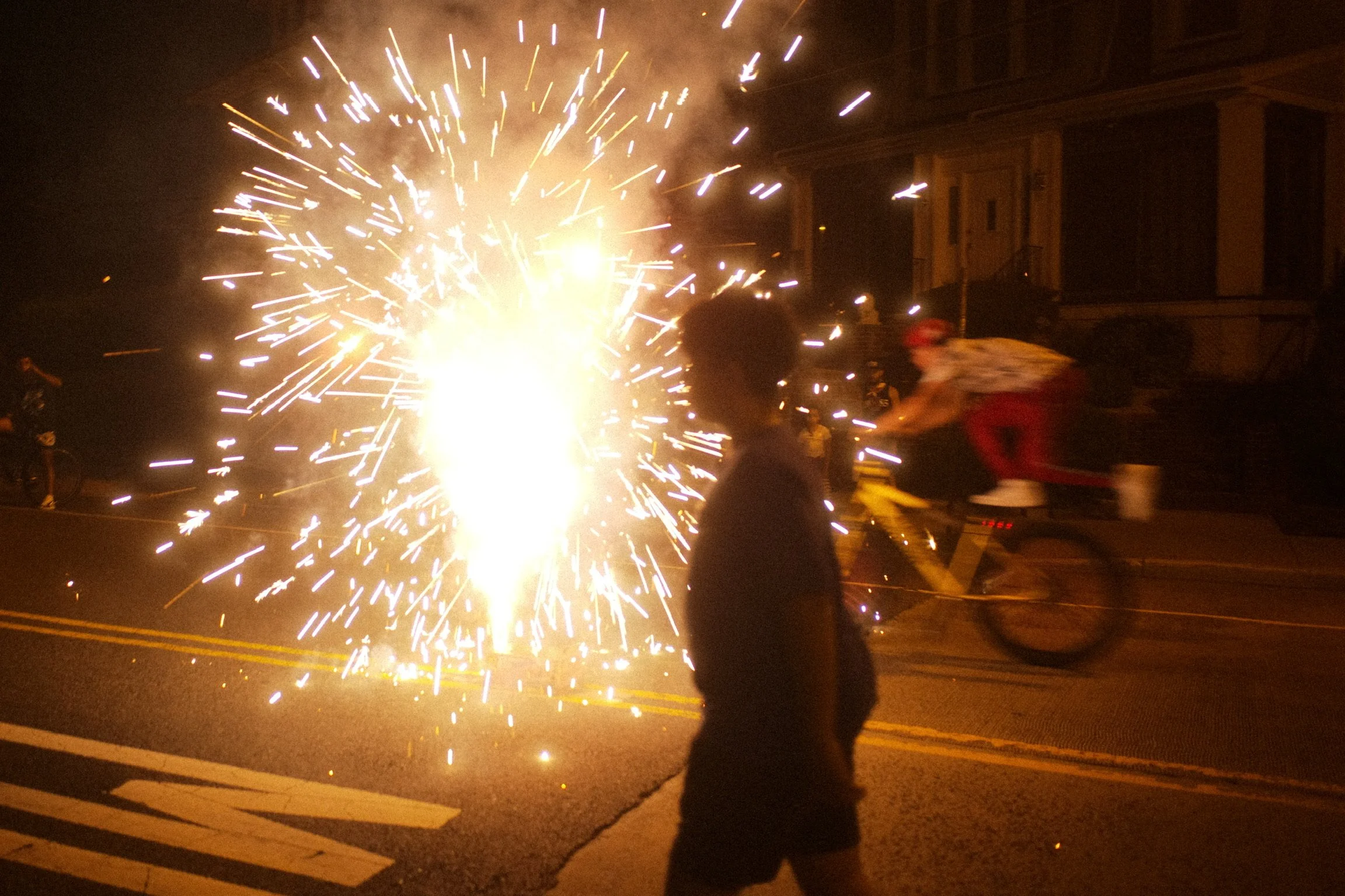 A person standing on a street at night near an outdoor firework or sparkler explosion, with a cyclist riding by in the background. The scene is illuminated by the bright sparks.
