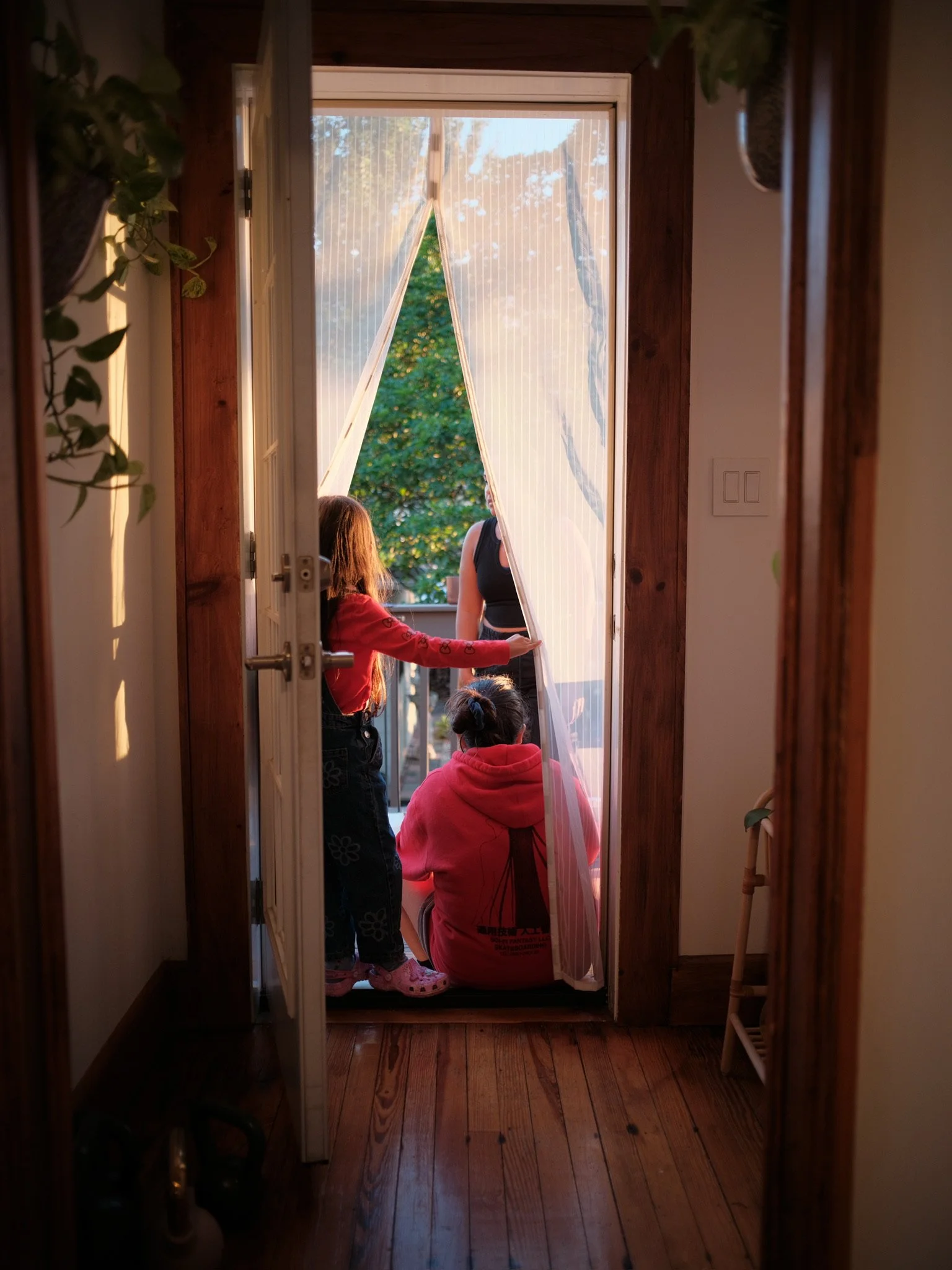 Three girls are gathered on a balcony, with the door open, as they look outside. One girl is standing, the other two are seated, with sunlight illuminating the scene and greenery in the background.