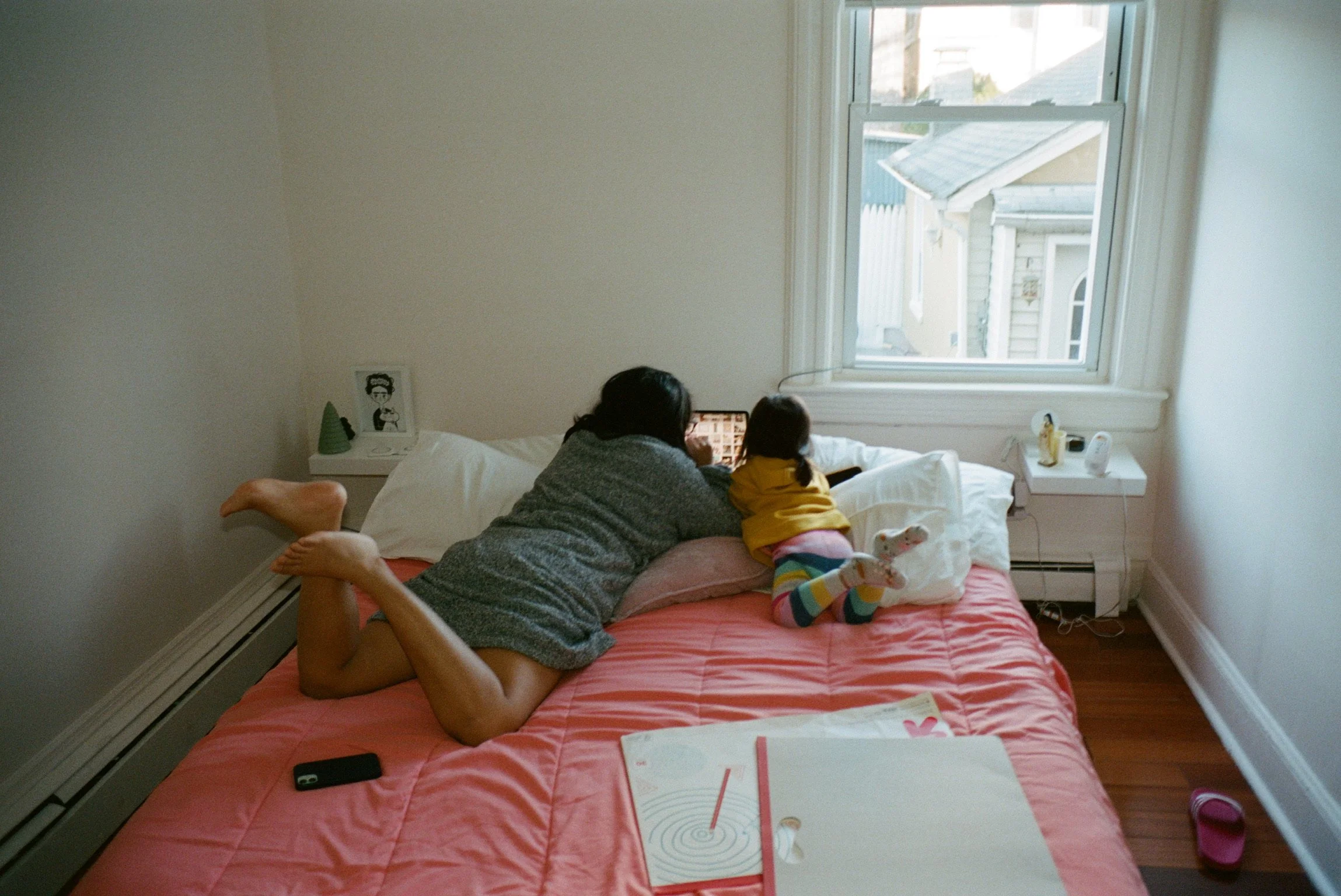 Person and child lying on bed looking at a tablet, with a window in the background showing a house and trees.