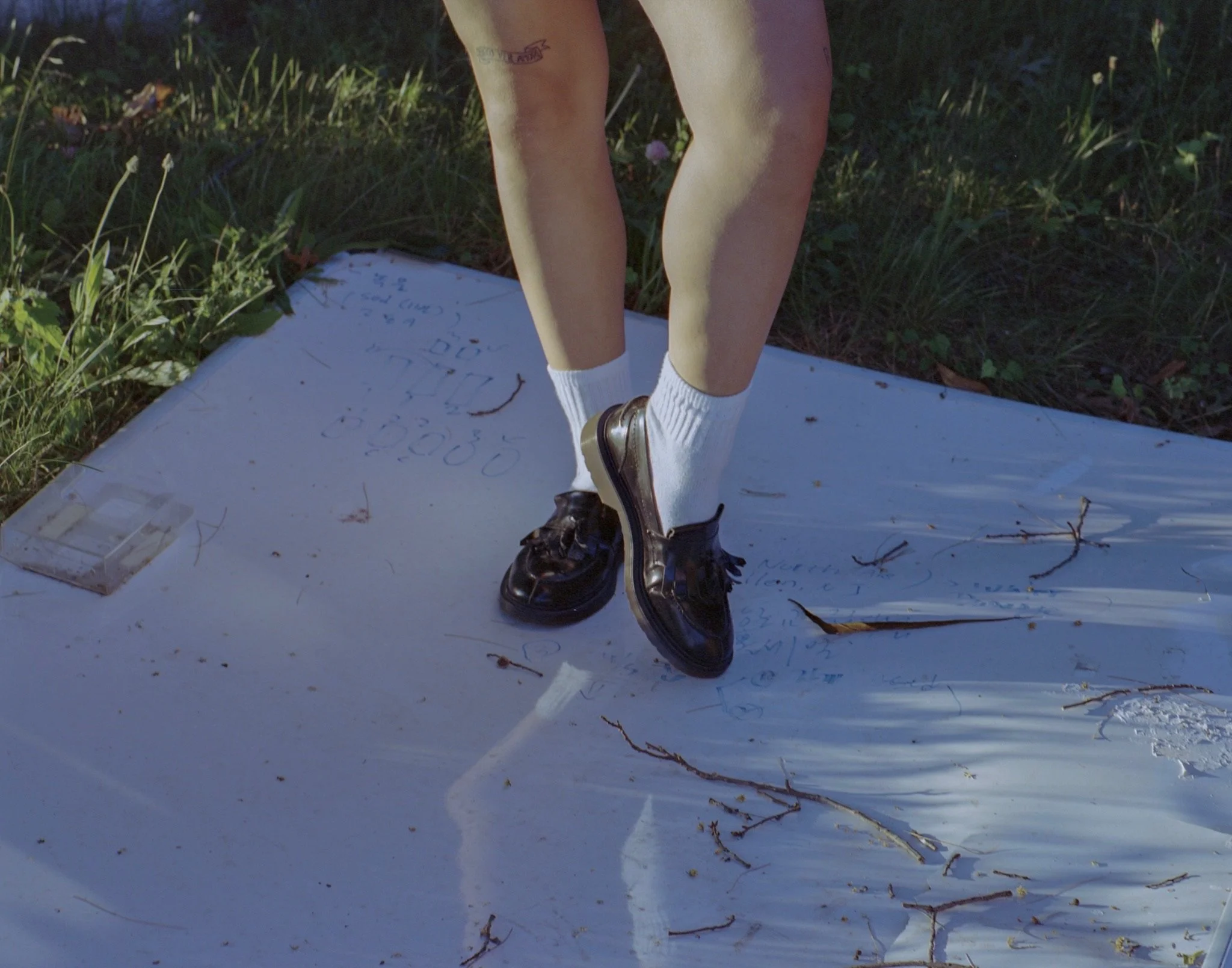 Close-up of a person's legs and feet standing on a white surface outdoors. The person is wearing white socks and black shoes, and has a tattoo on their right thigh. The background includes grass and some small plants.