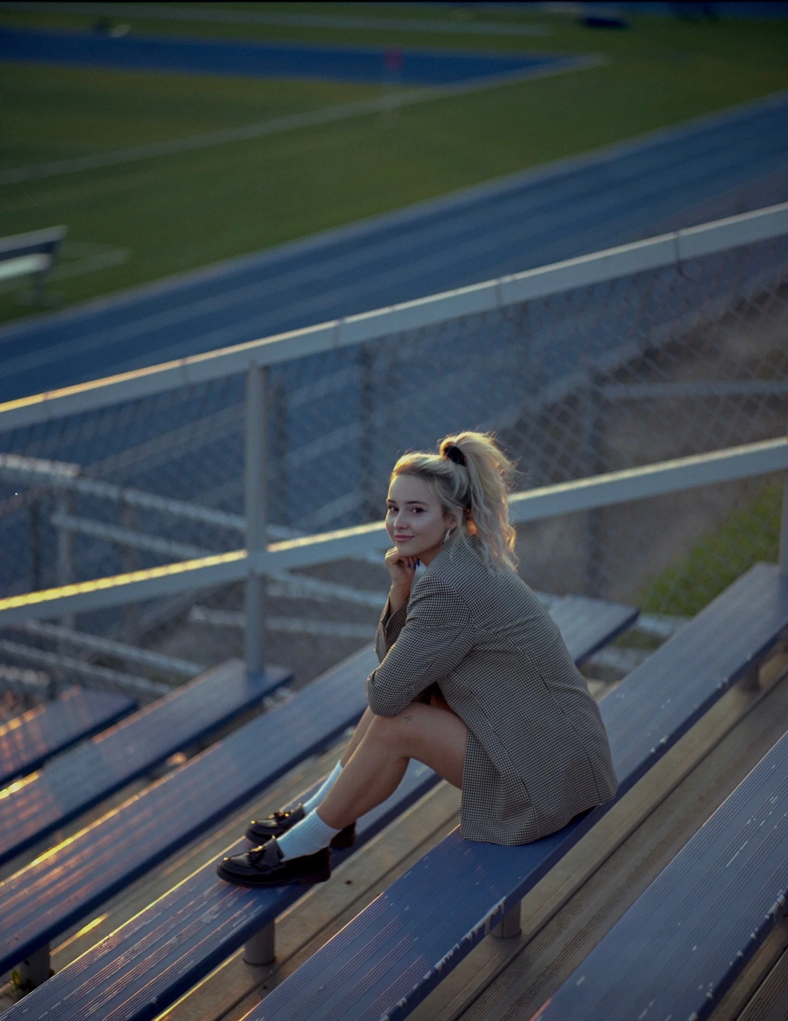 A young woman with blonde hair in a high ponytail sitting alone on empty sports bleachers at a track stadium during sunset, wearing a checkered blazer, white socks, and black loafers, smiling and looking at the camera.