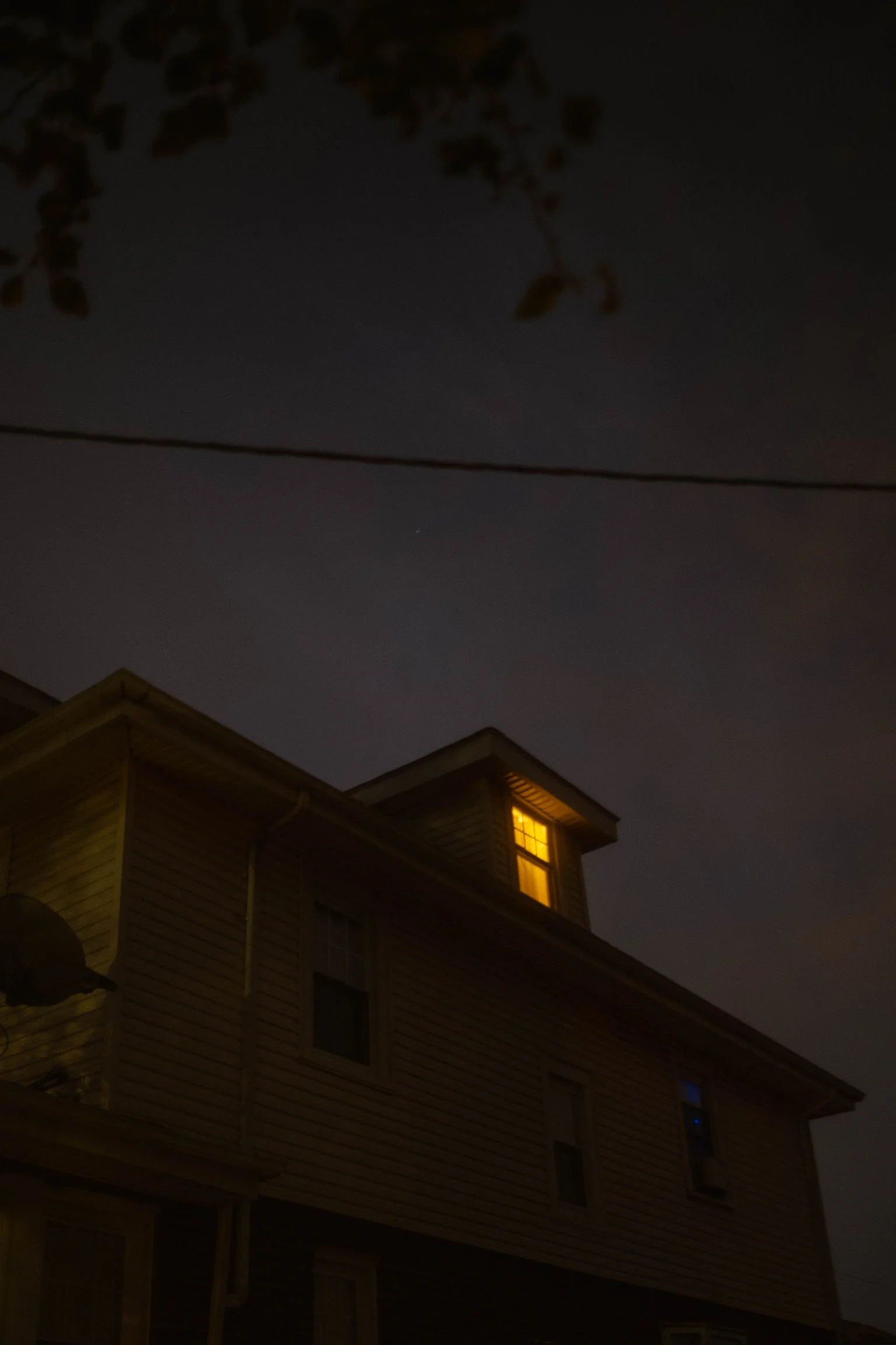 Nighttime photo of a house with a lit window on the upper floor, dark sky, and faint outlines of tree branches and a wire overhead.