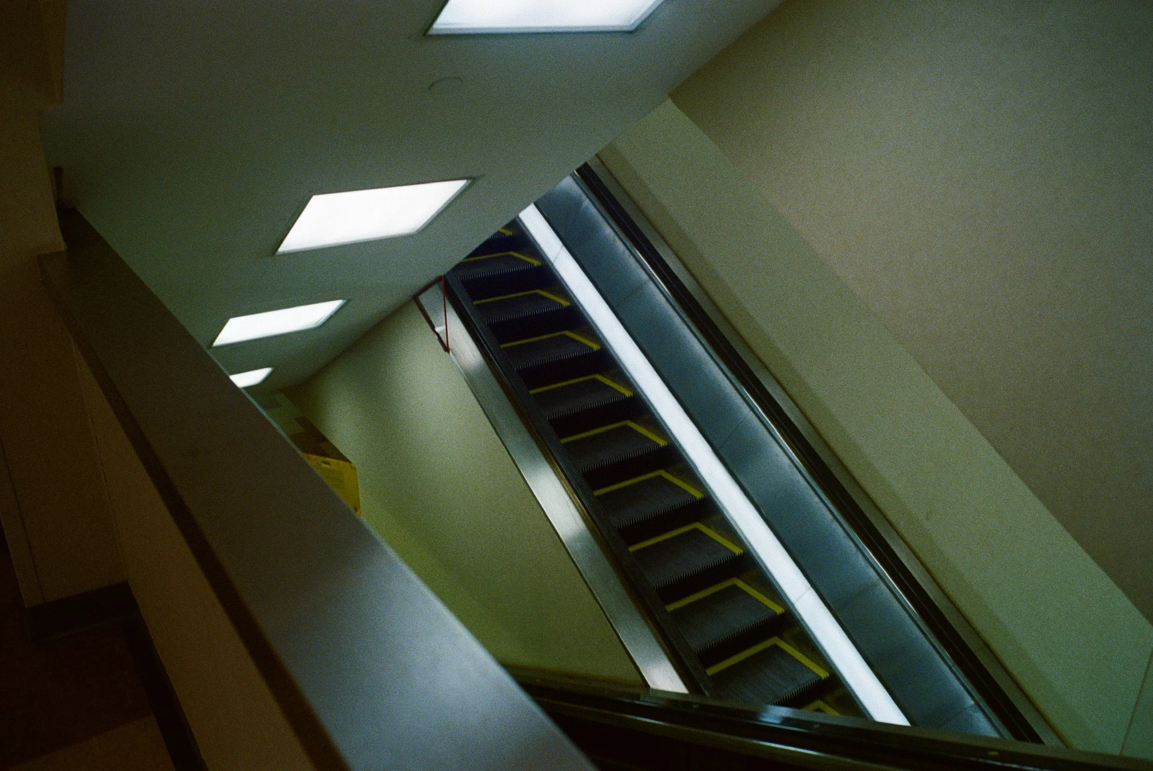 Escalator descending in an indoor setting, with a view toward the ceiling and walls, illuminated by rectangular ceiling lights.