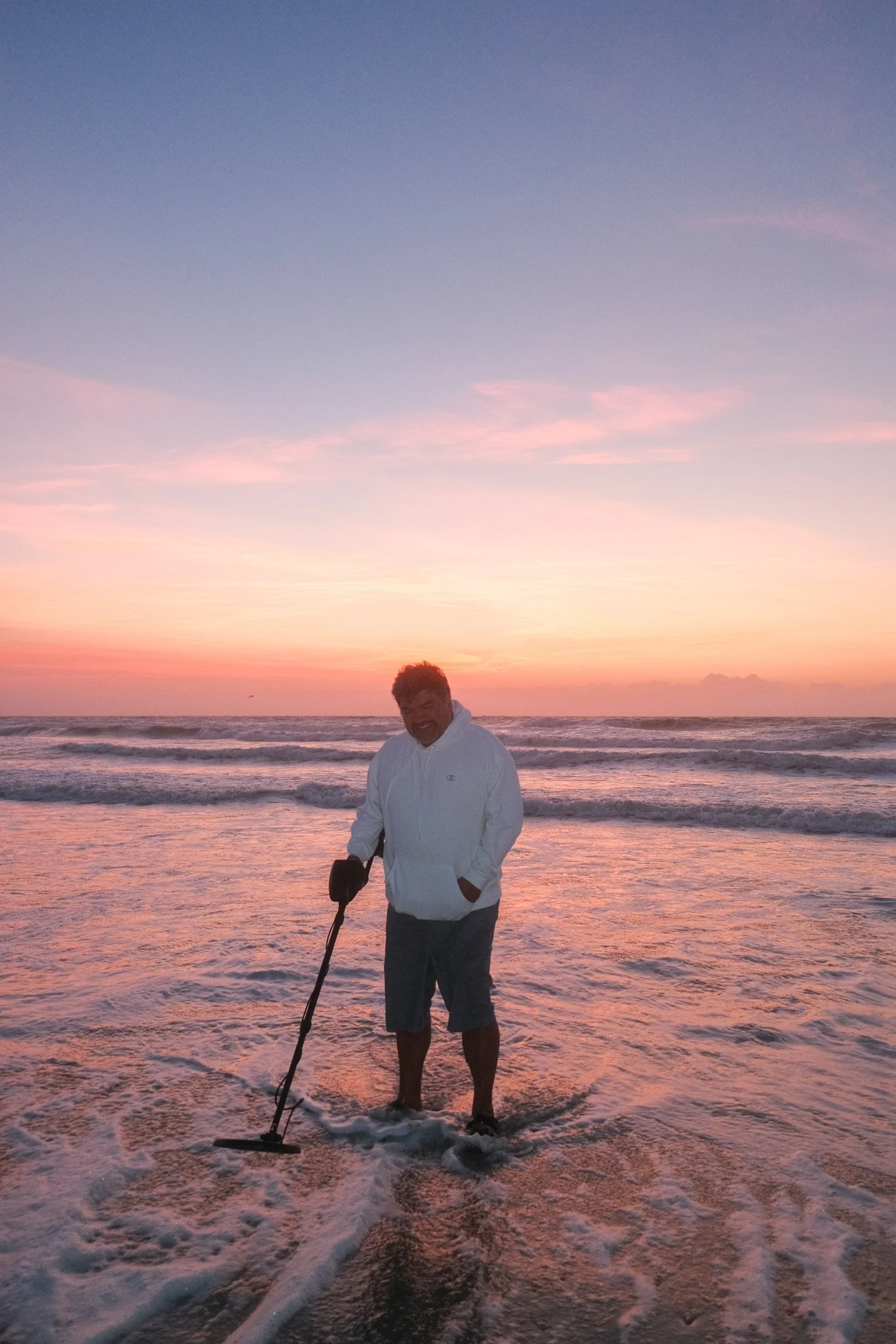 A man standing in the ocean at sunset while using a metal detector, wearing a white hoodie and shorts, smiling, with pink and purple sky in the background.