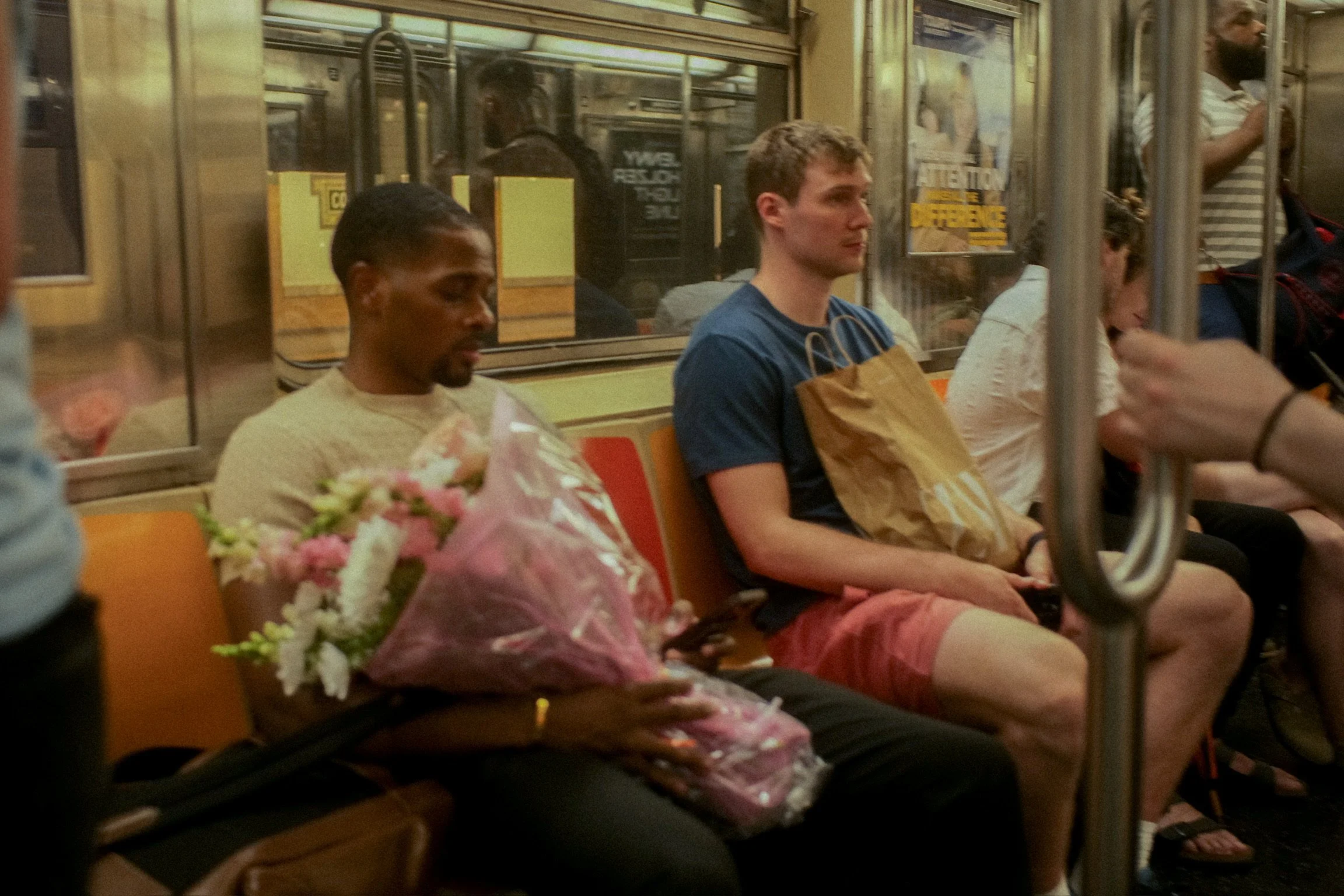 A man holding a bouquet of flowers on a subway train, sitting next to another man with a brown paper shopping bag, with others seated around.
