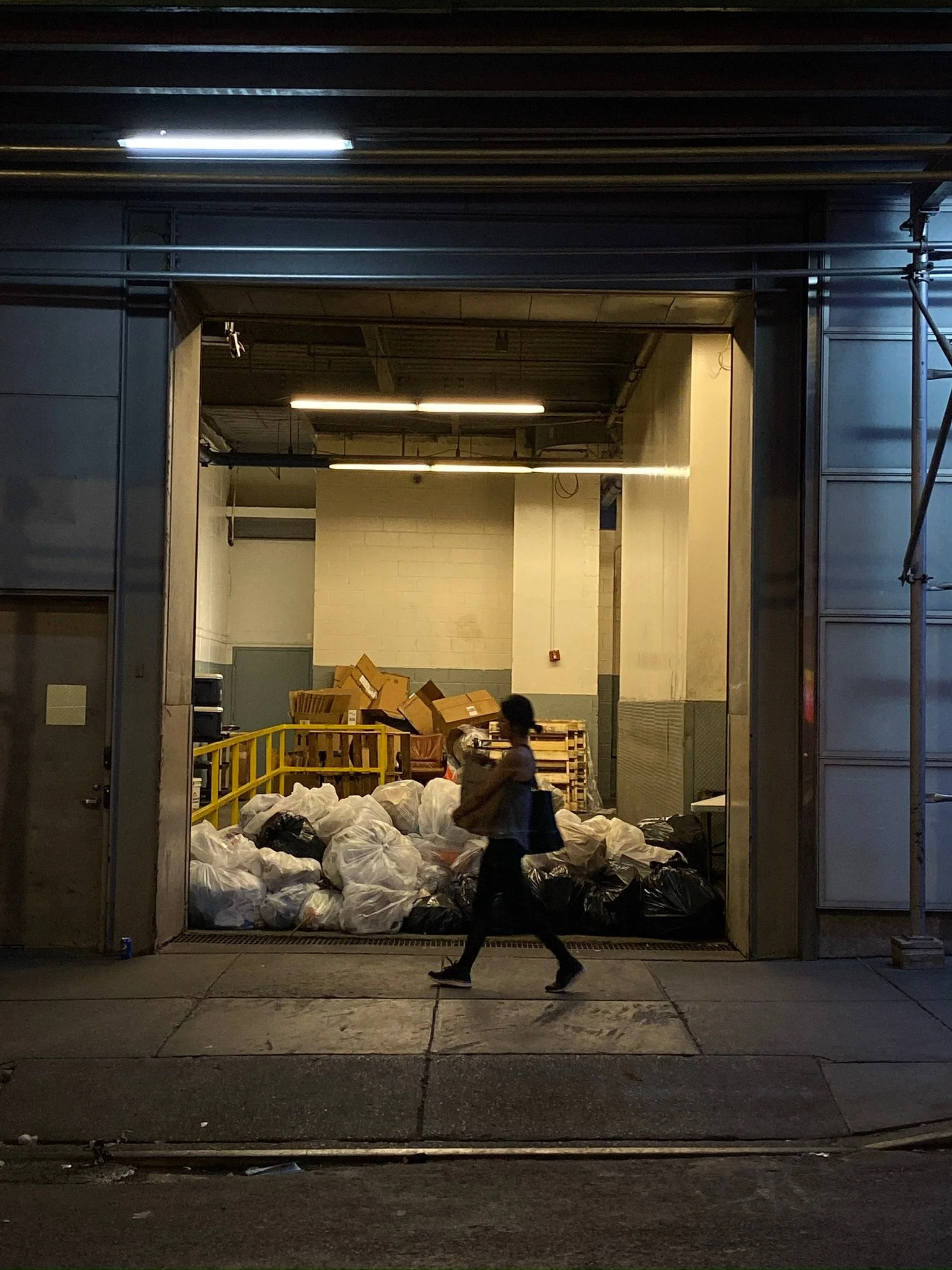 A person carrying bags walks past a large industrial trash collection area with piled garbage bags and cardboard boxes inside a warehouse