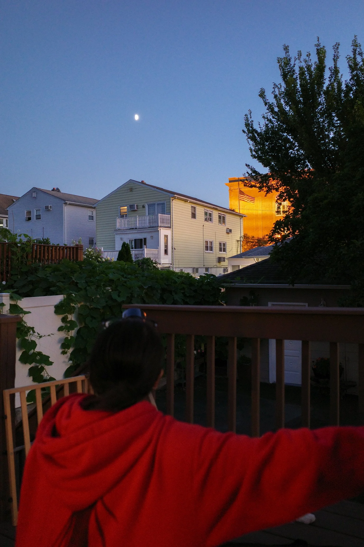 A person in a red hoodie sitting on a wooden deck, looking at houses and a water tower with an American flag, under a blue sky with a visible moon.
