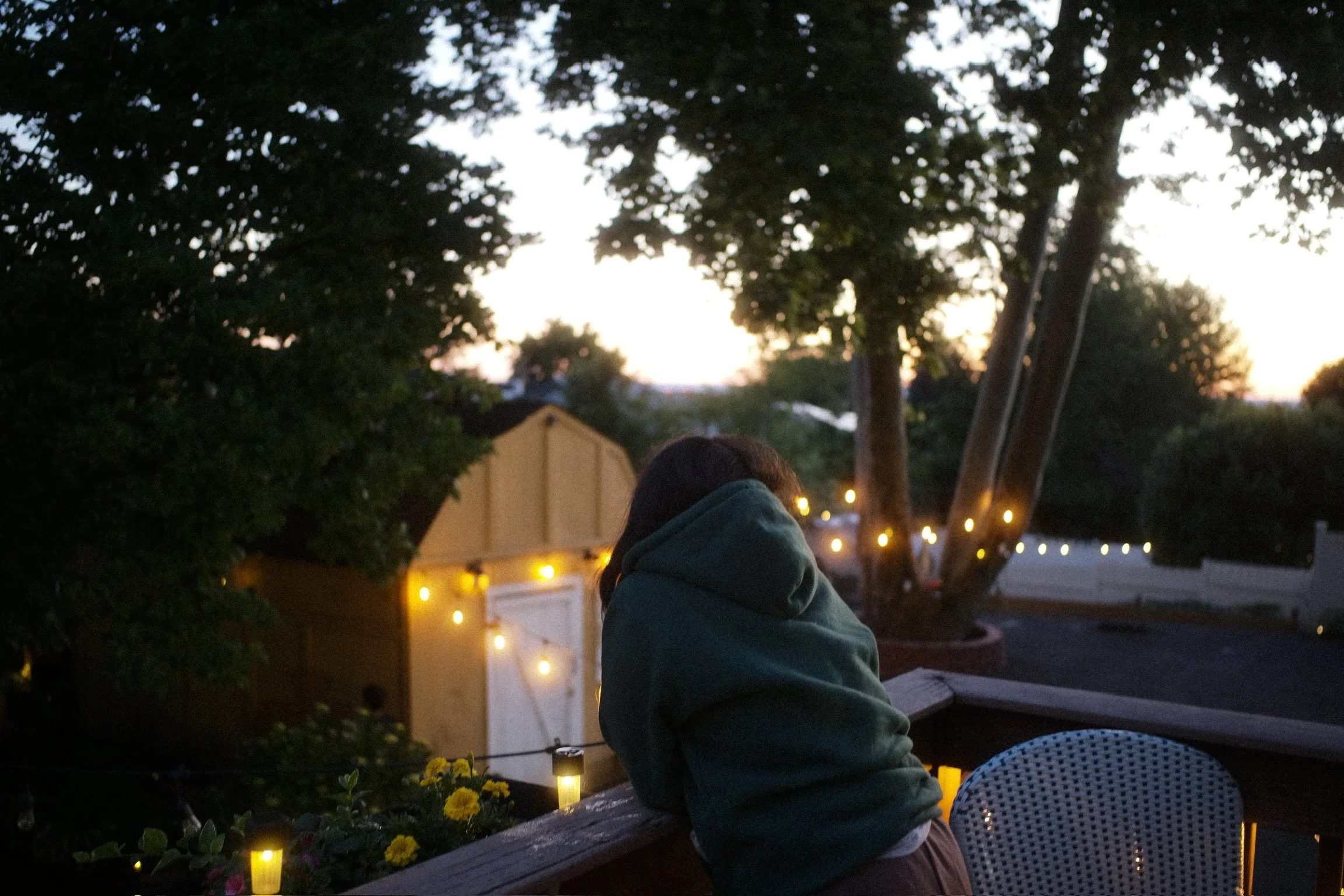 Person with a hoodie leaning on a wooden railing on a deck during sunset, with trees and houses in the background decorated with string lights.