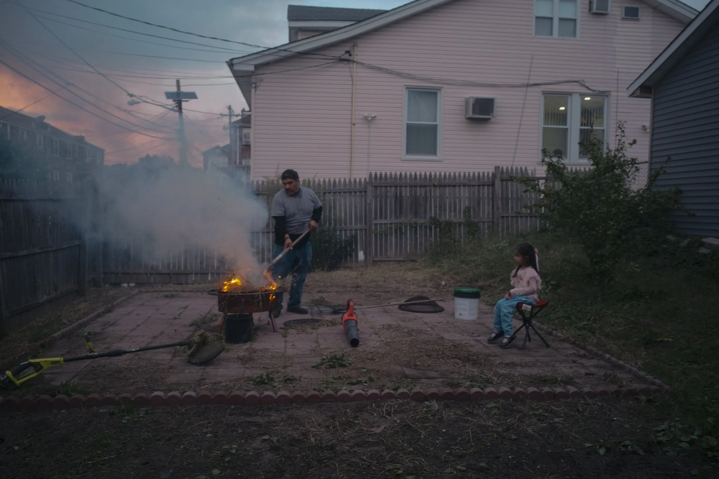 A man and a young girl sitting outside in a backyard at dusk, with the man stoking a small fire in a portable grill, producing smoke and small flames. The girl is seated nearby on a folding chair, observing. The yard has a paved area, a fence, some h