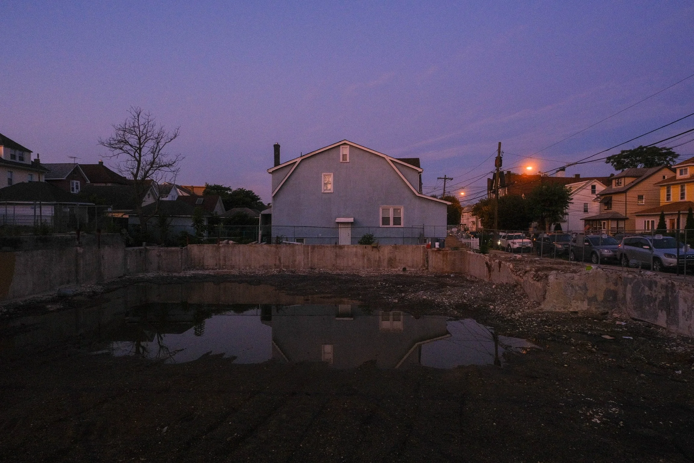 Residential neighborhood during dusk with a central empty lot containing a small pool of water, surrounded by houses, power lines, and streetlights.