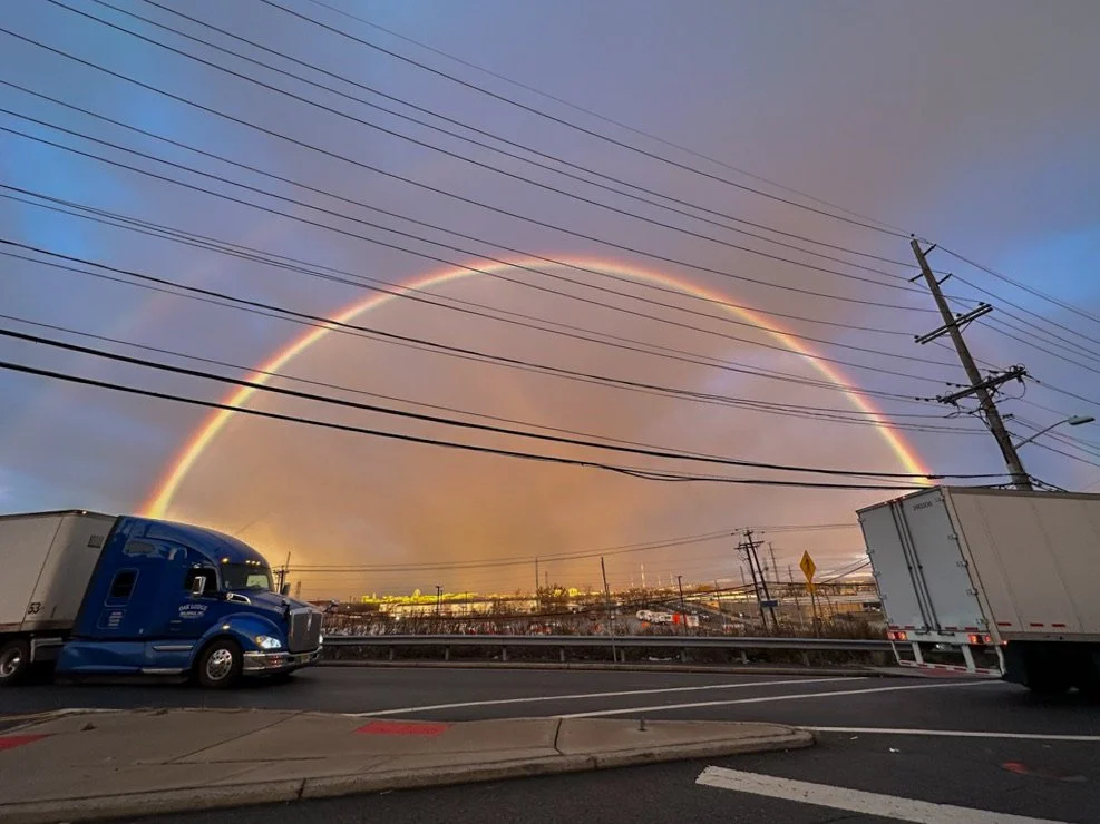 A rainbow arching across the sky over a highway with trucks and power lines, during a sunset or sunrise.