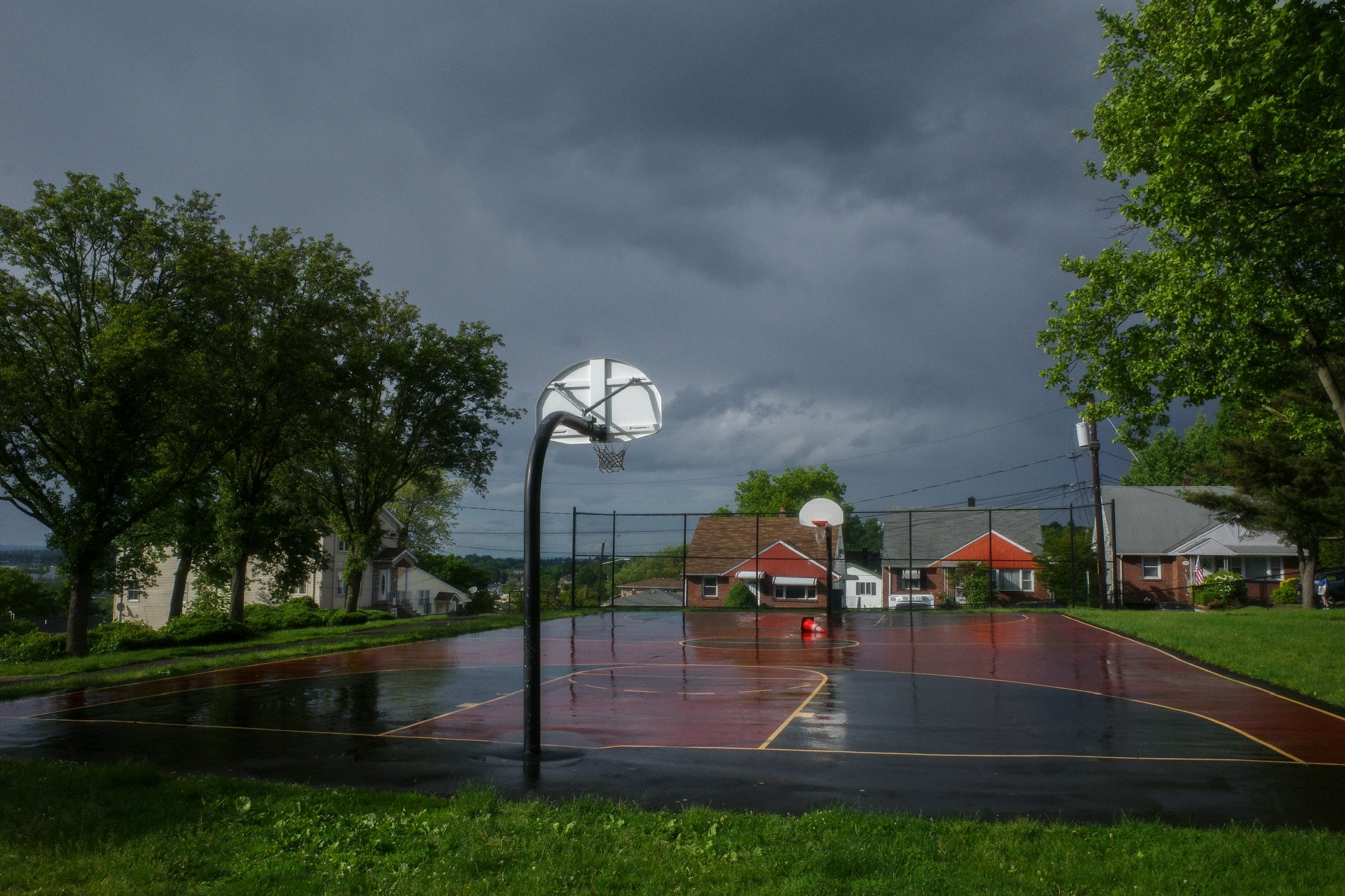 Empty outdoor basketball court on a rainy day with wet surface, dark storm clouds overhead, and houses in the background.