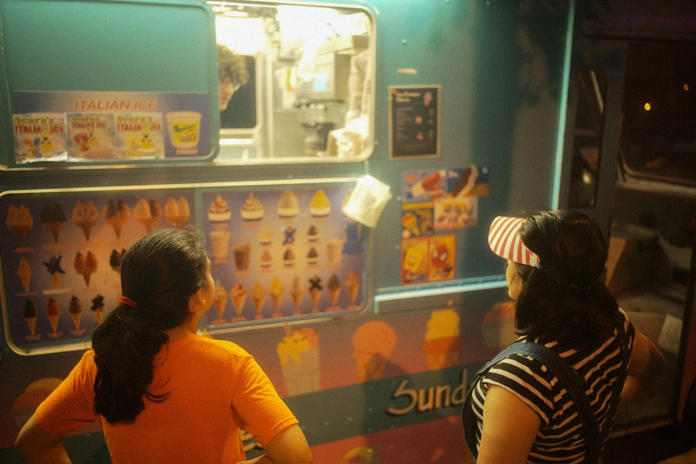 Two women standing in front of an ice cream vending machine at night, looking at the selection of ice creams inside.