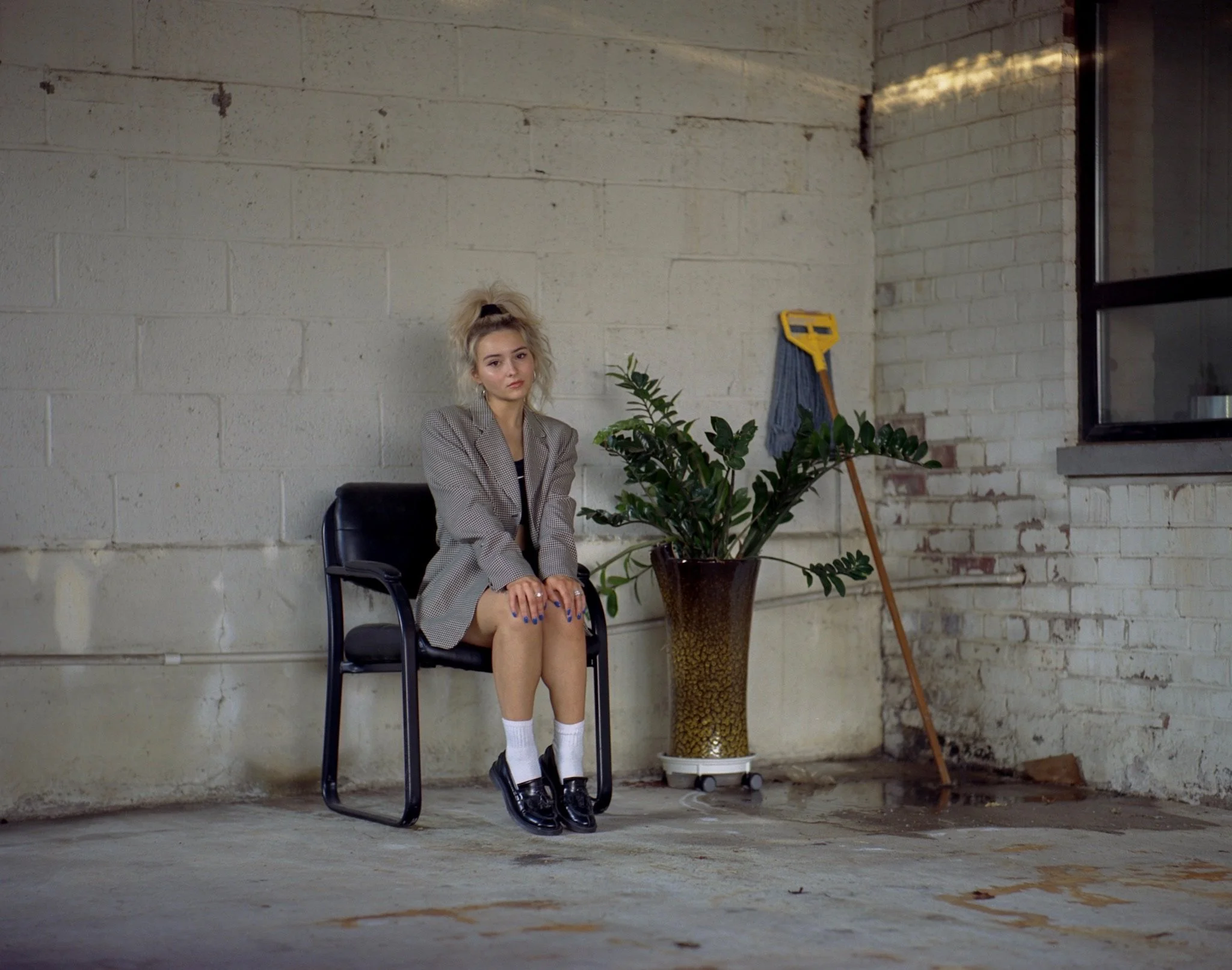 A young woman with blonde hair tied in a high ponytail, wearing a checkered blazer, white socks, and black shoes, sitting alone on a black chair in a room with white brick walls and concrete floor. There is a large potted plant, a yellow mop, and a w