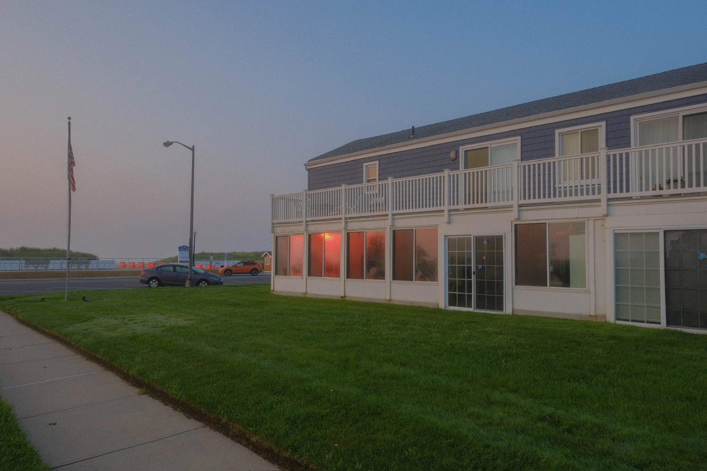 A two-story building with a blue exterior and white balcony railings, located near a grassy area, with a sidewalk, cars, streetlights, and a flagpole with an American flag in the background, during dusk.