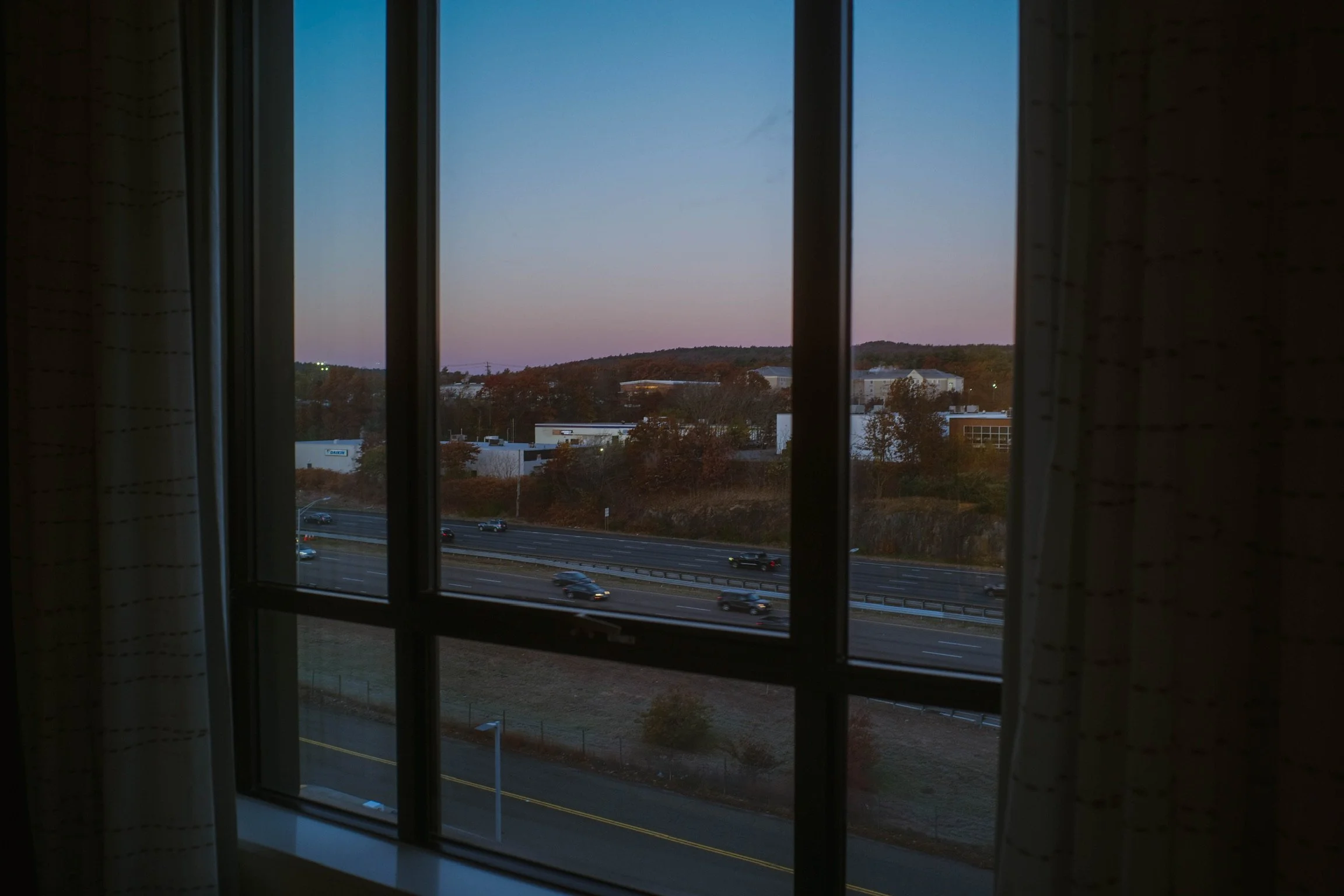 View through a window showing a highway with cars, trees, and distant buildings during dusk or dawn.