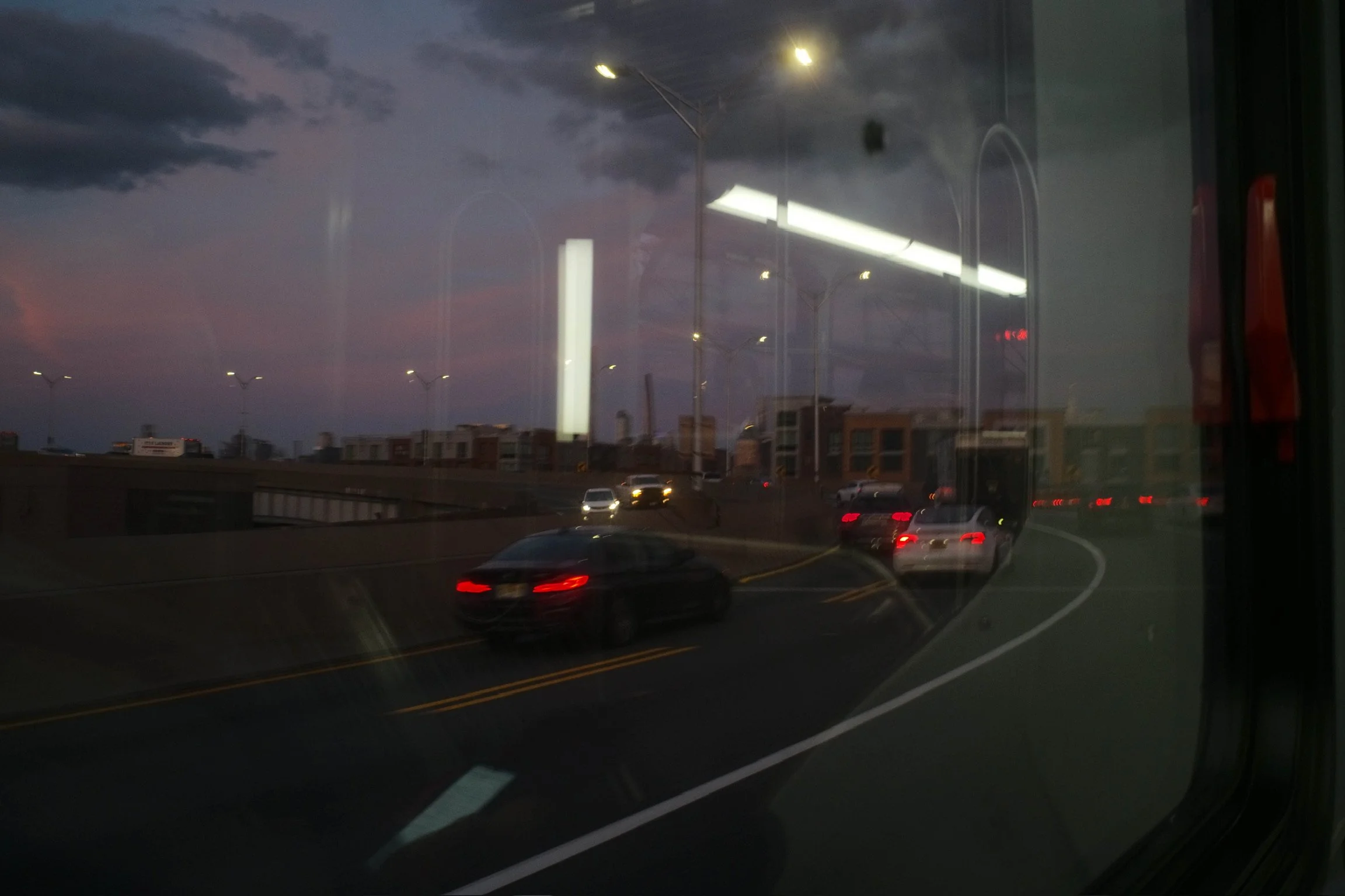 View of cars on a highway at dusk seen through a window, with reflections of interior lights and parts of the bus or train interior.