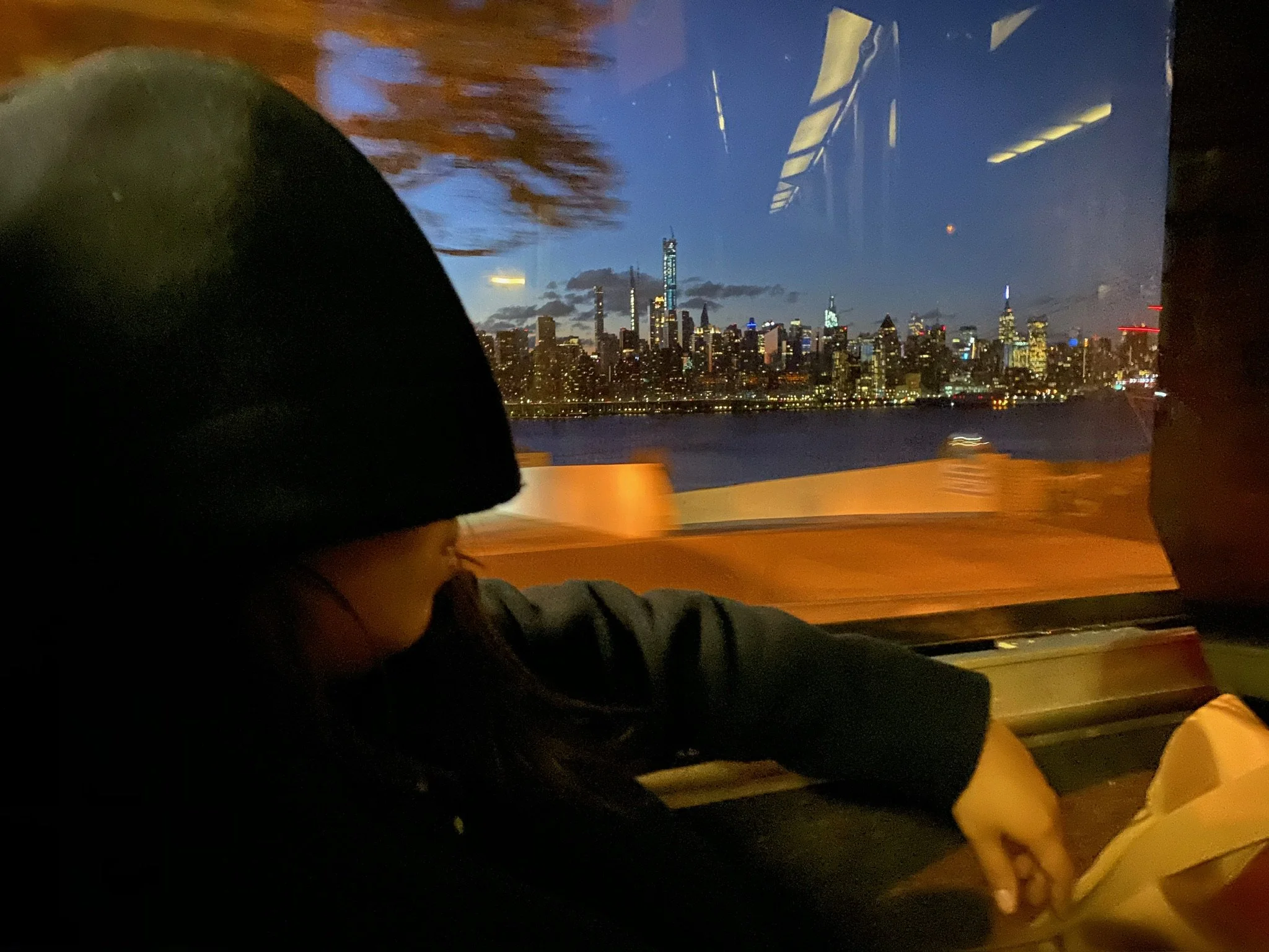 A woman wearing a hoodie sitting on a bus with a city skyline at dusk in the background.
